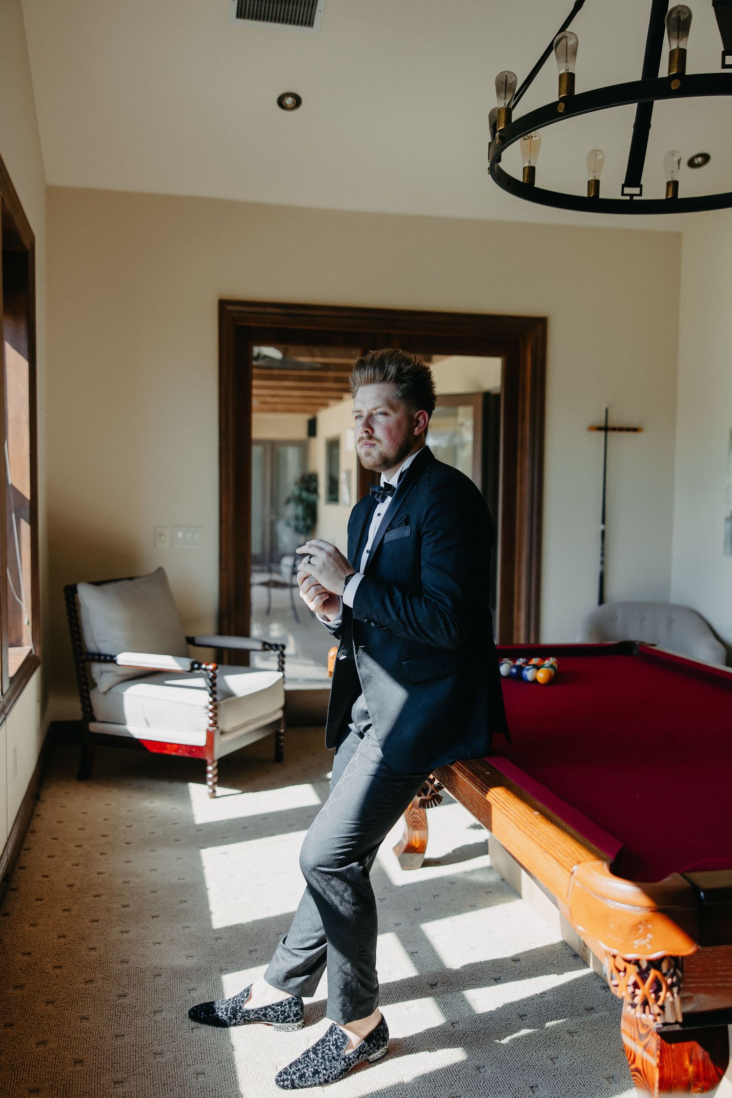Groom in classic black tuxedo standing in sunlit room with pool table.