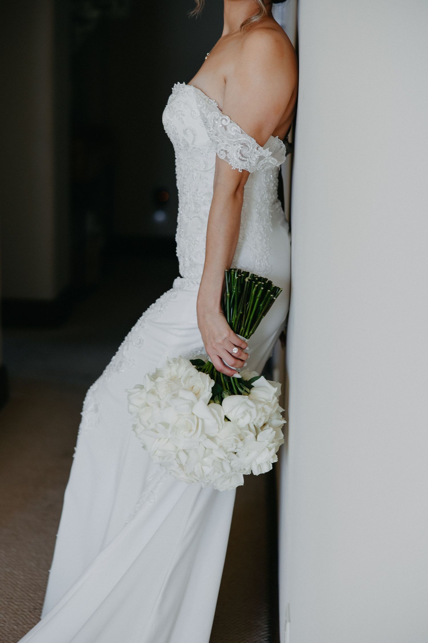 Side view of bride in fitted lace wedding dress holding white bouquet against hallway wall.