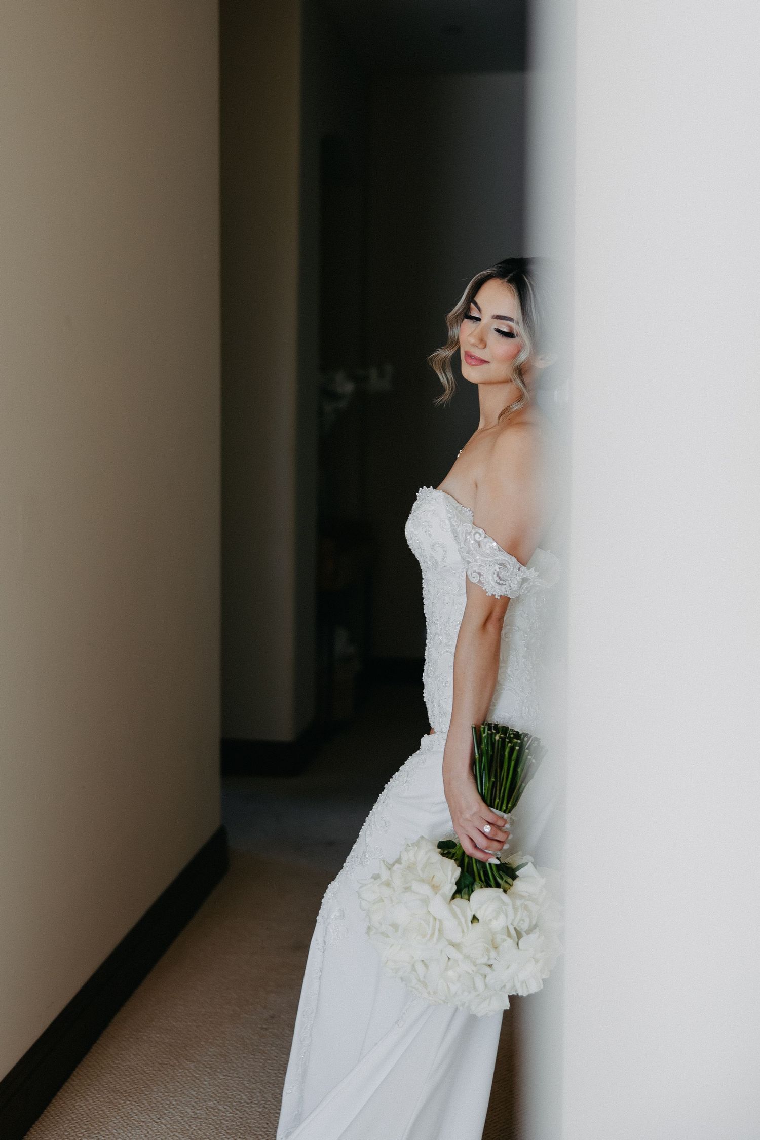 Bride in white off-shoulder wedding gown holds a bouquet while standing against a wall.