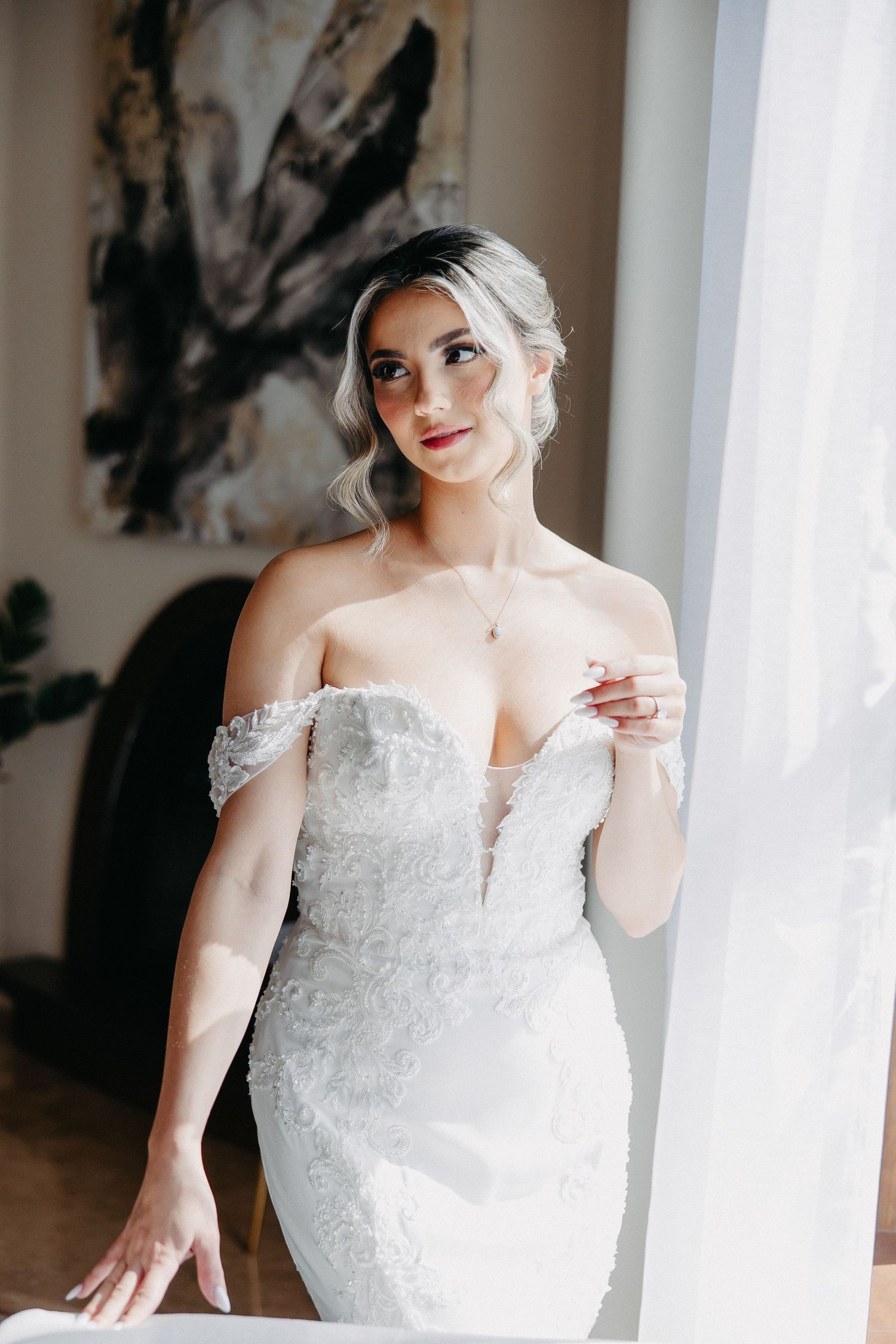 Elegant bride in fitted lace wedding dress poses by window with abstract art in background.
