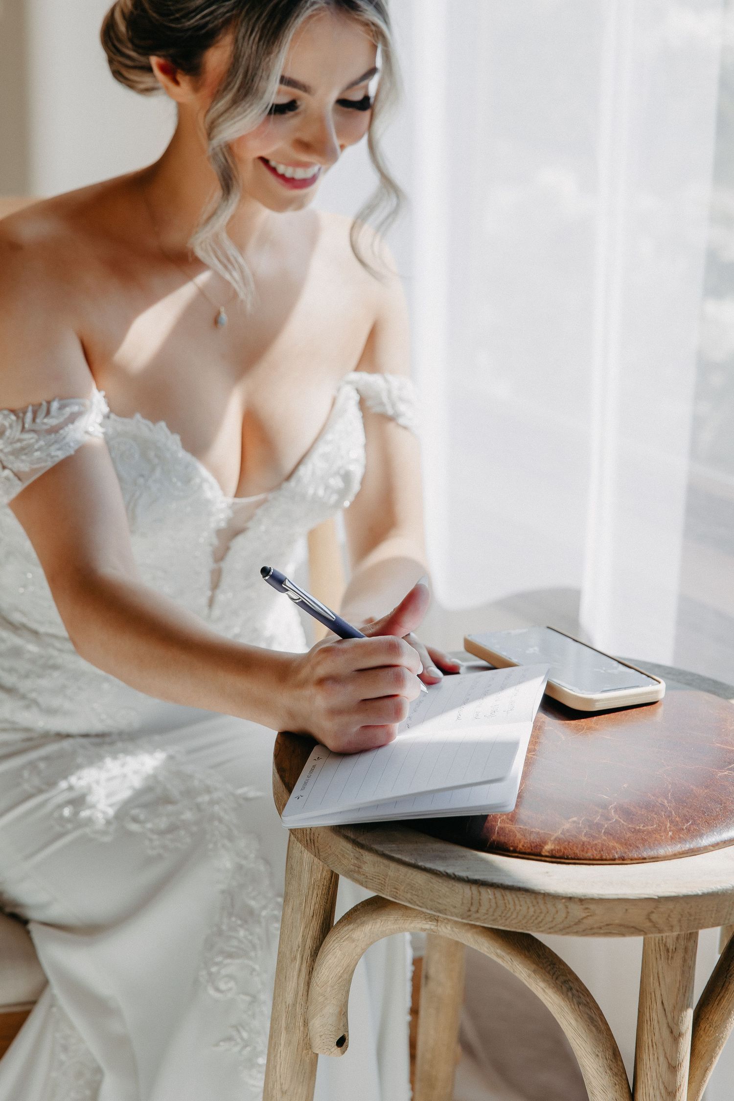 Bride in white lace dress writes in notebook while sitting at small wooden table.