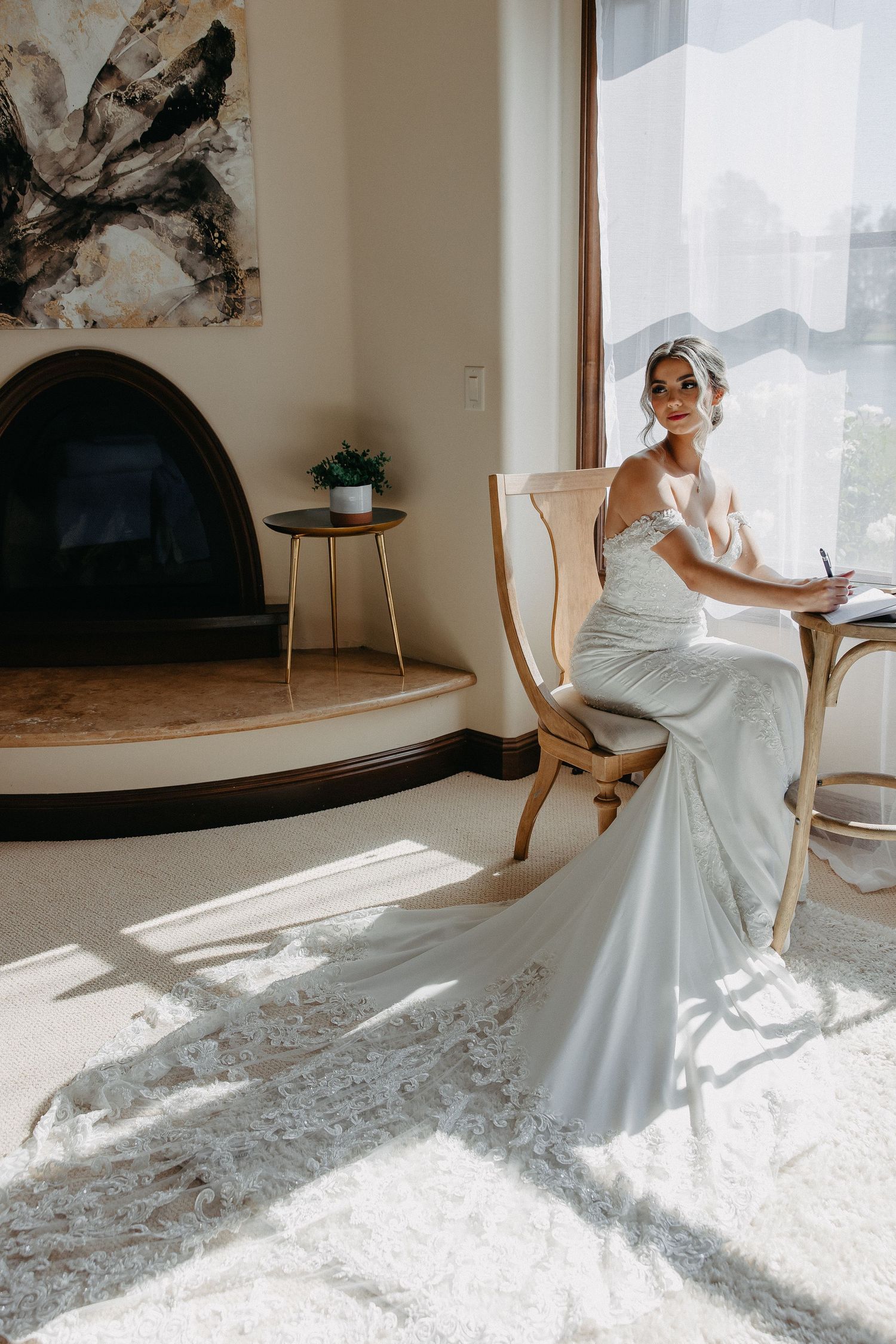 Bride in elegant white dress sits in vintage chair near fireplace with natural light streaming in.