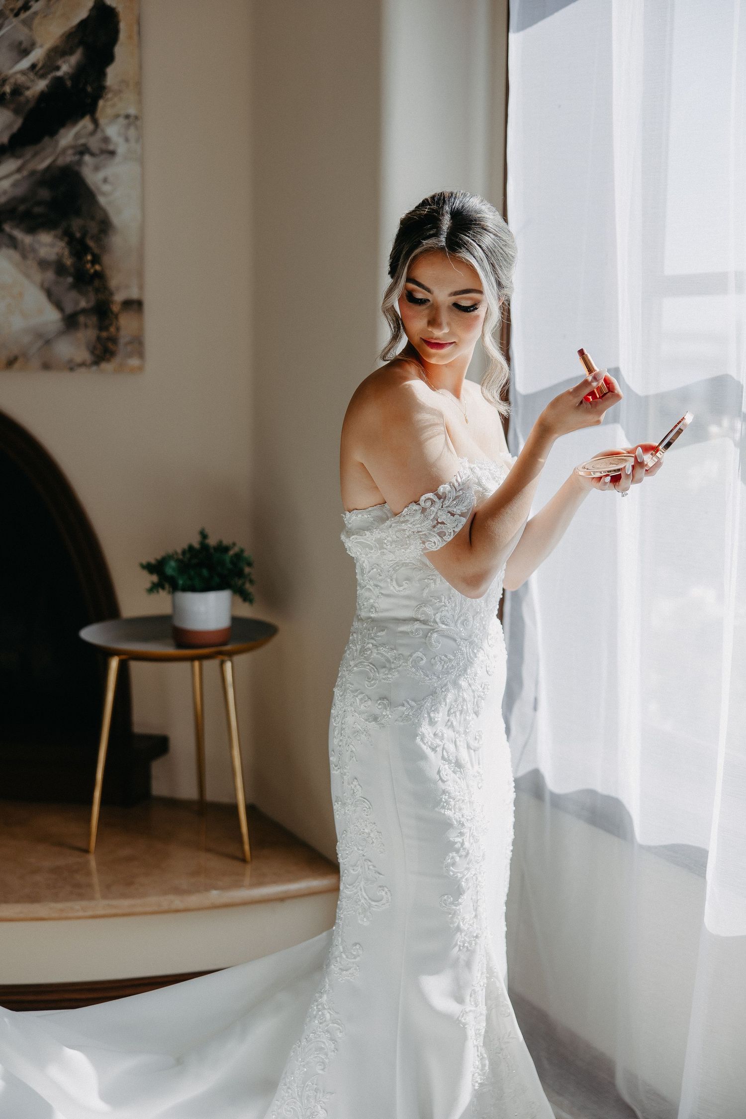 Bride in form-fitting lace gown checks phone near decorative side table with potted plant.
