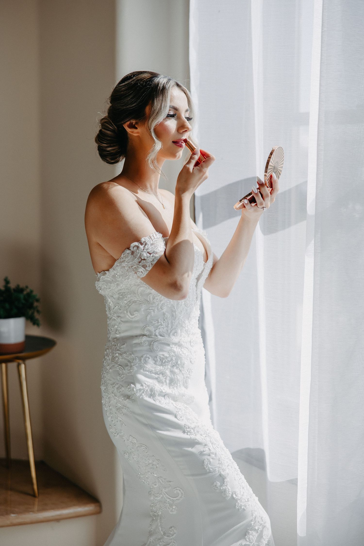 Bride in lace wedding dress applies lipstick while looking in handheld mirror near window.