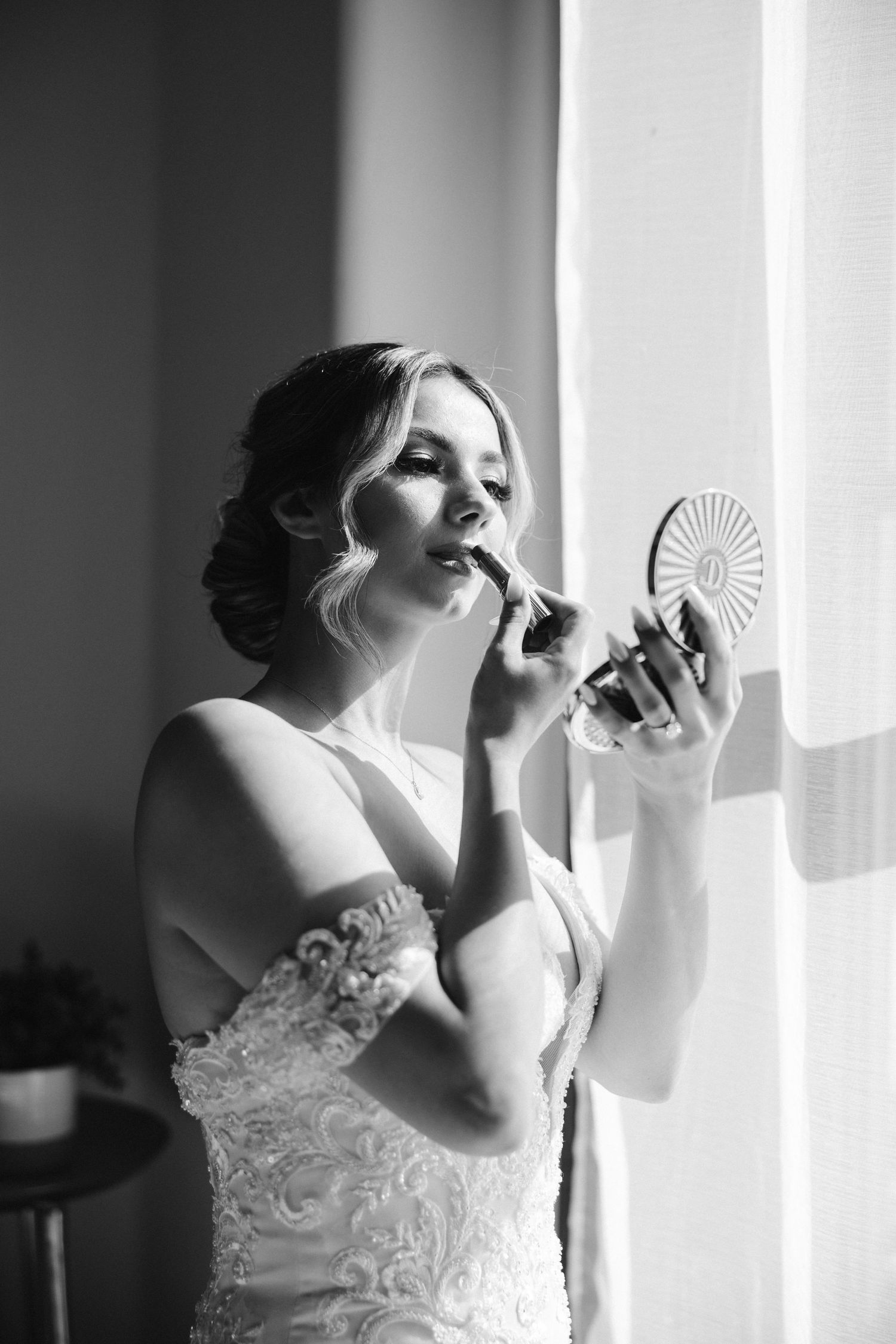 Black and white portrait of bride applying lipstick using compact mirror by window.