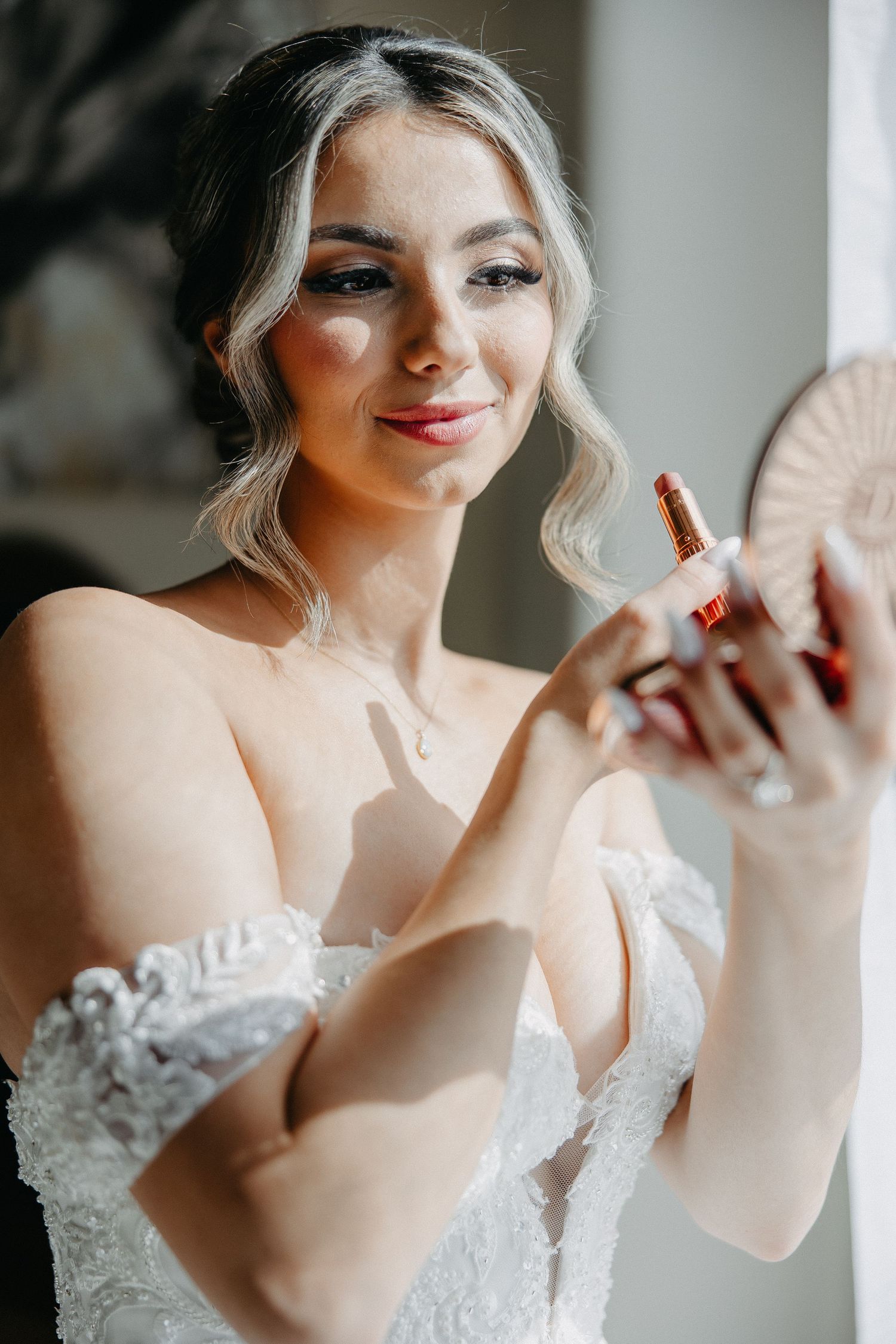 Bride in a white lace wedding dress applies lip color while getting ready for her ceremony.