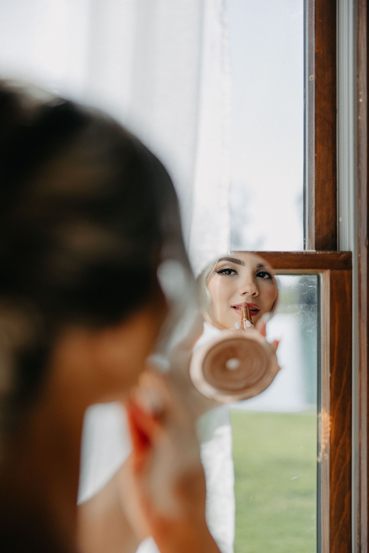 Close-up shot of a bride applying makeup in a circular mirror by a window.