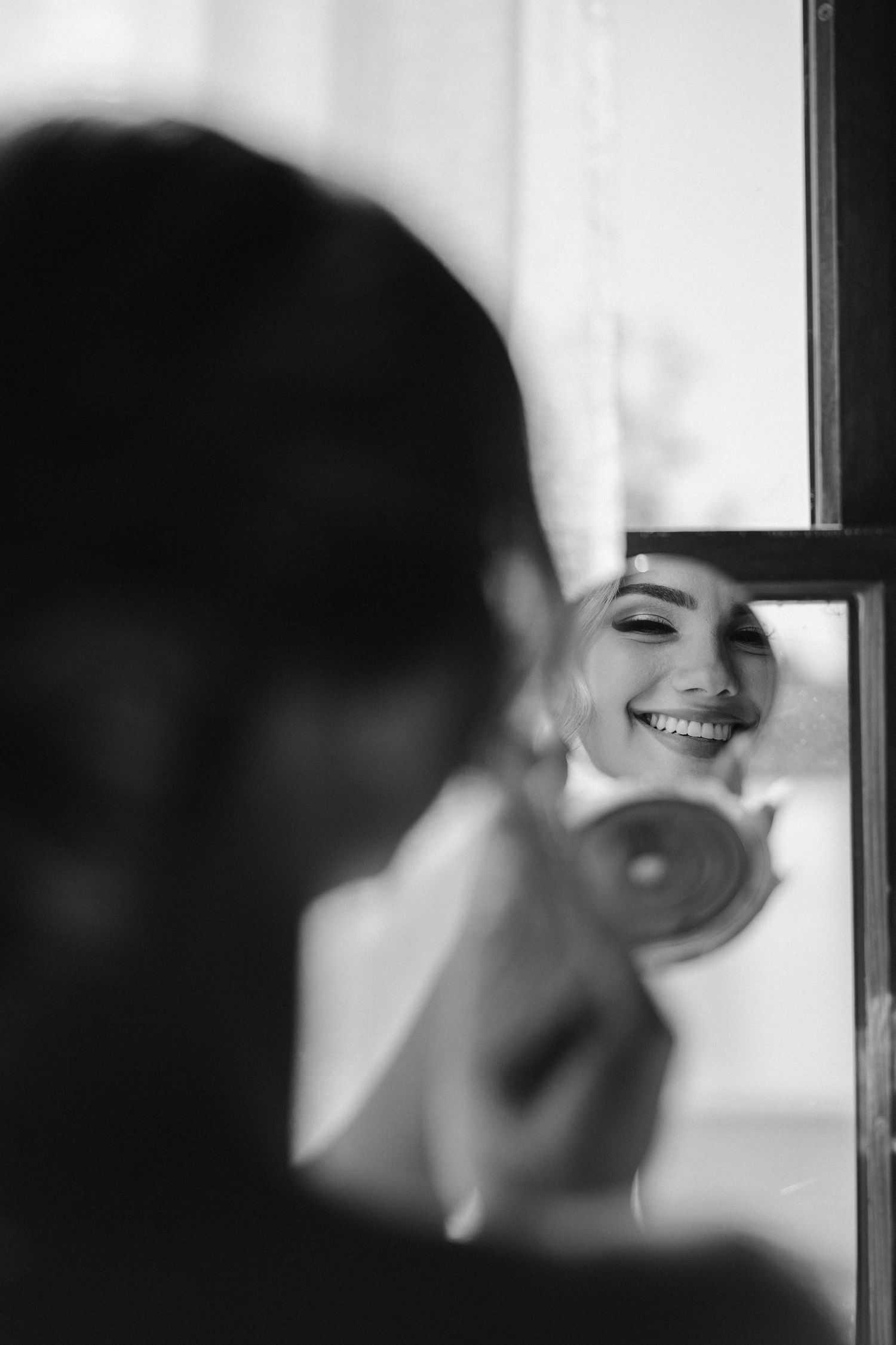 Black and white reflection of a bride smiling in a mirror during wedding preparations.