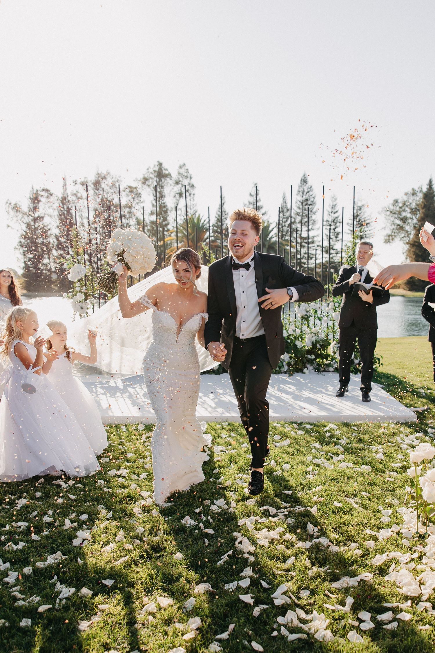 Wedding party celebrates on lawn with confetti and balloons against backdrop of tall pine trees.