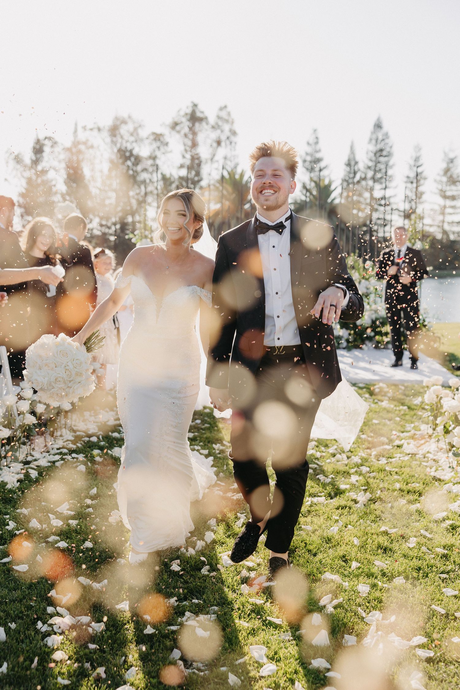Joyful newlyweds run through confetti shower during outdoor forest wedding celebration.