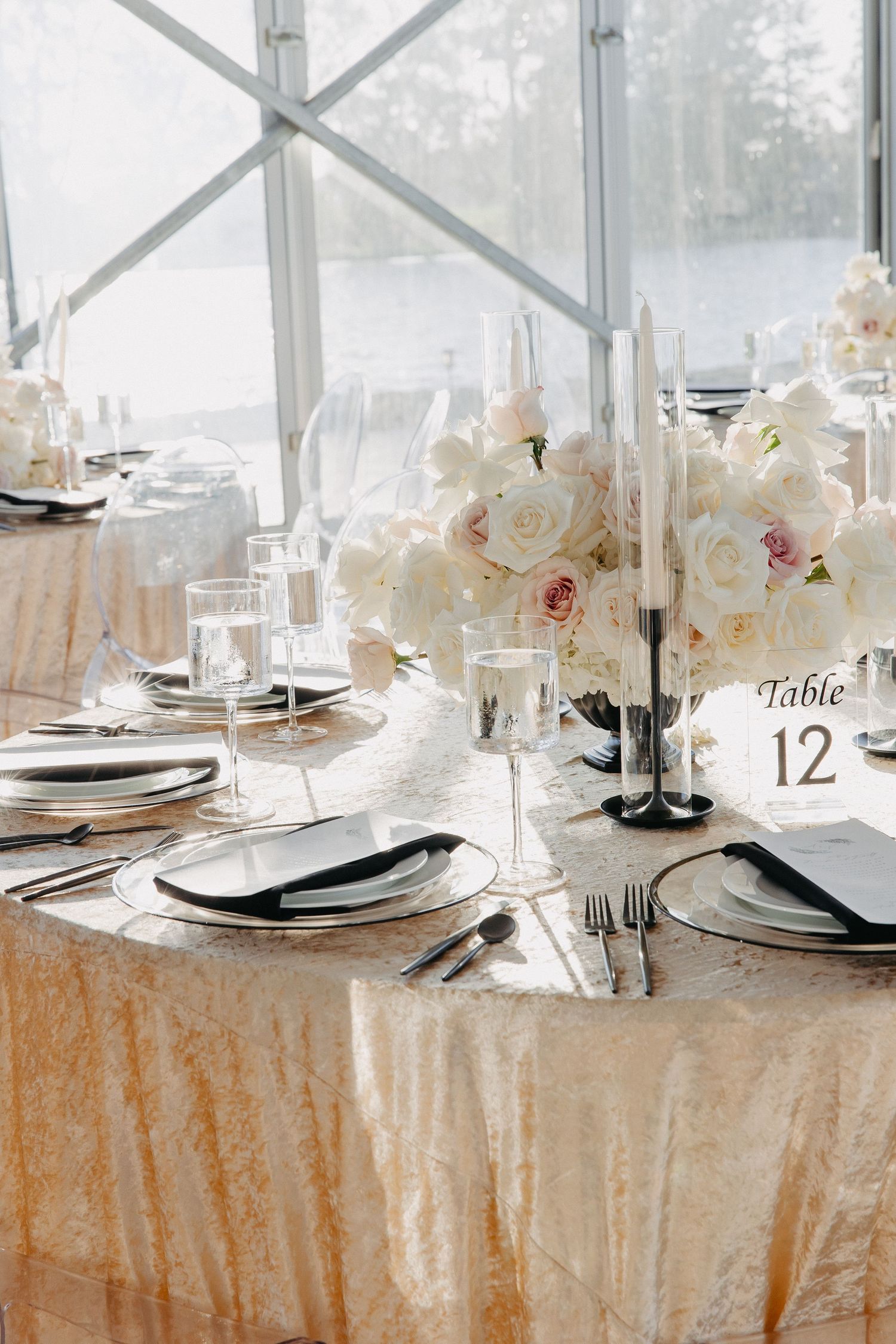 Elegant wedding table setting with white roses and champagne tablecloth in a sunlit tent venue.