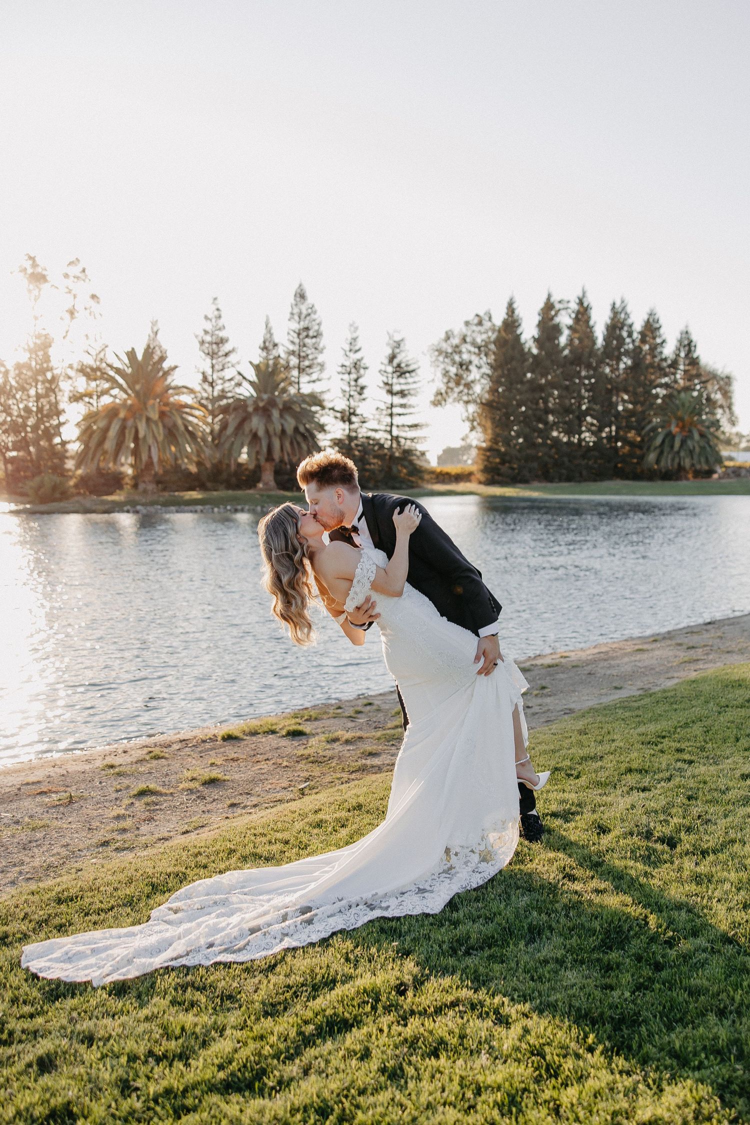 Wedding couple sharing romantic dip pose by lake at sunset with palm trees and evergreens in background.