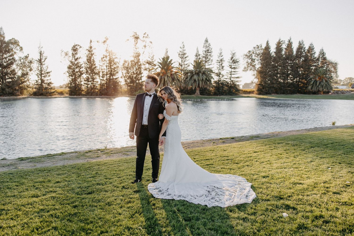 Newlyweds pose together on grassy lakeside with flowing white dress and evergreen trees at sunset.