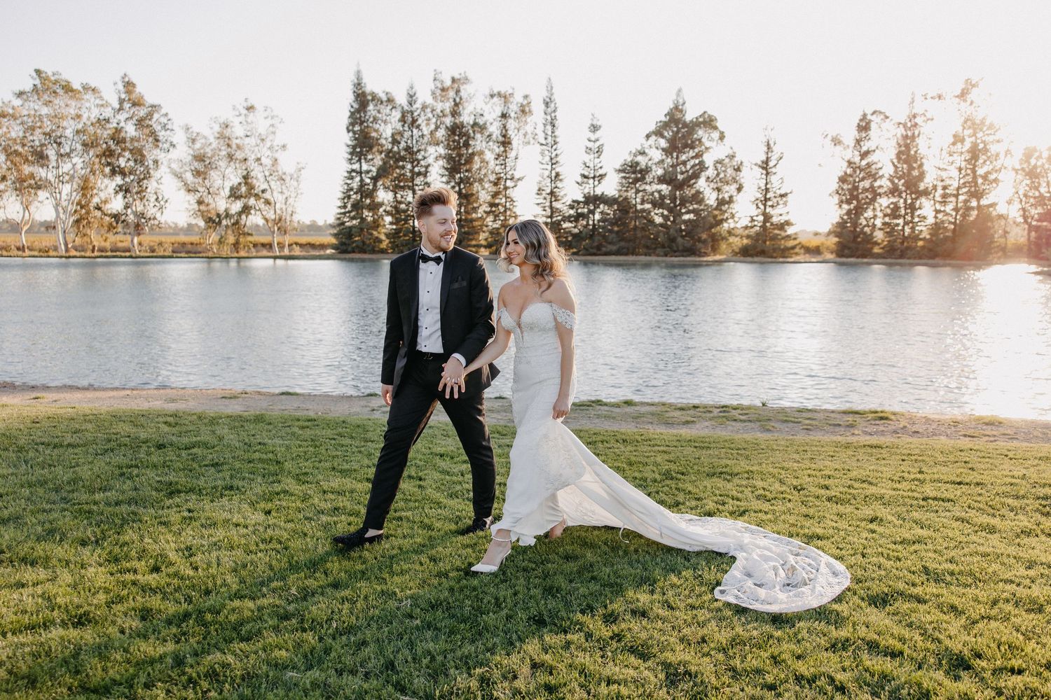 Wedding couple dancing playfully on grass by waterfront with long train of white dress trailing behind.