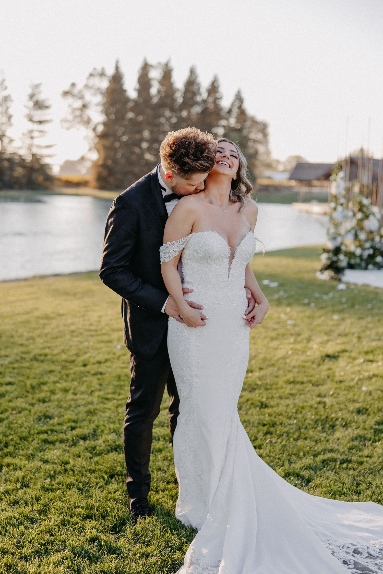 Intimate moment between newlyweds by lake with fitted off-shoulder wedding gown and formal attire.