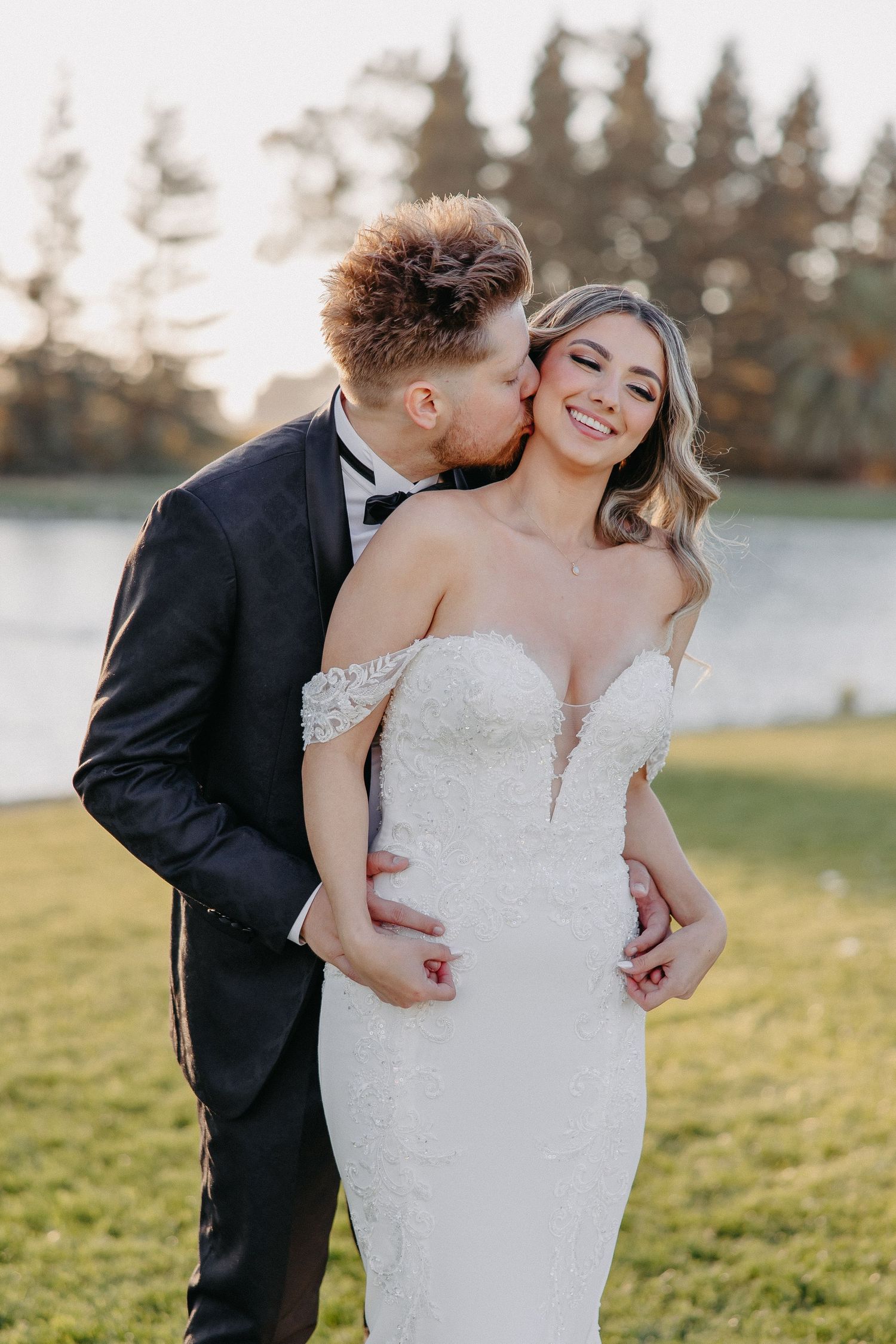 Tender wedding moment captured lakeside with fitted white bridal gown and black tuxedo.