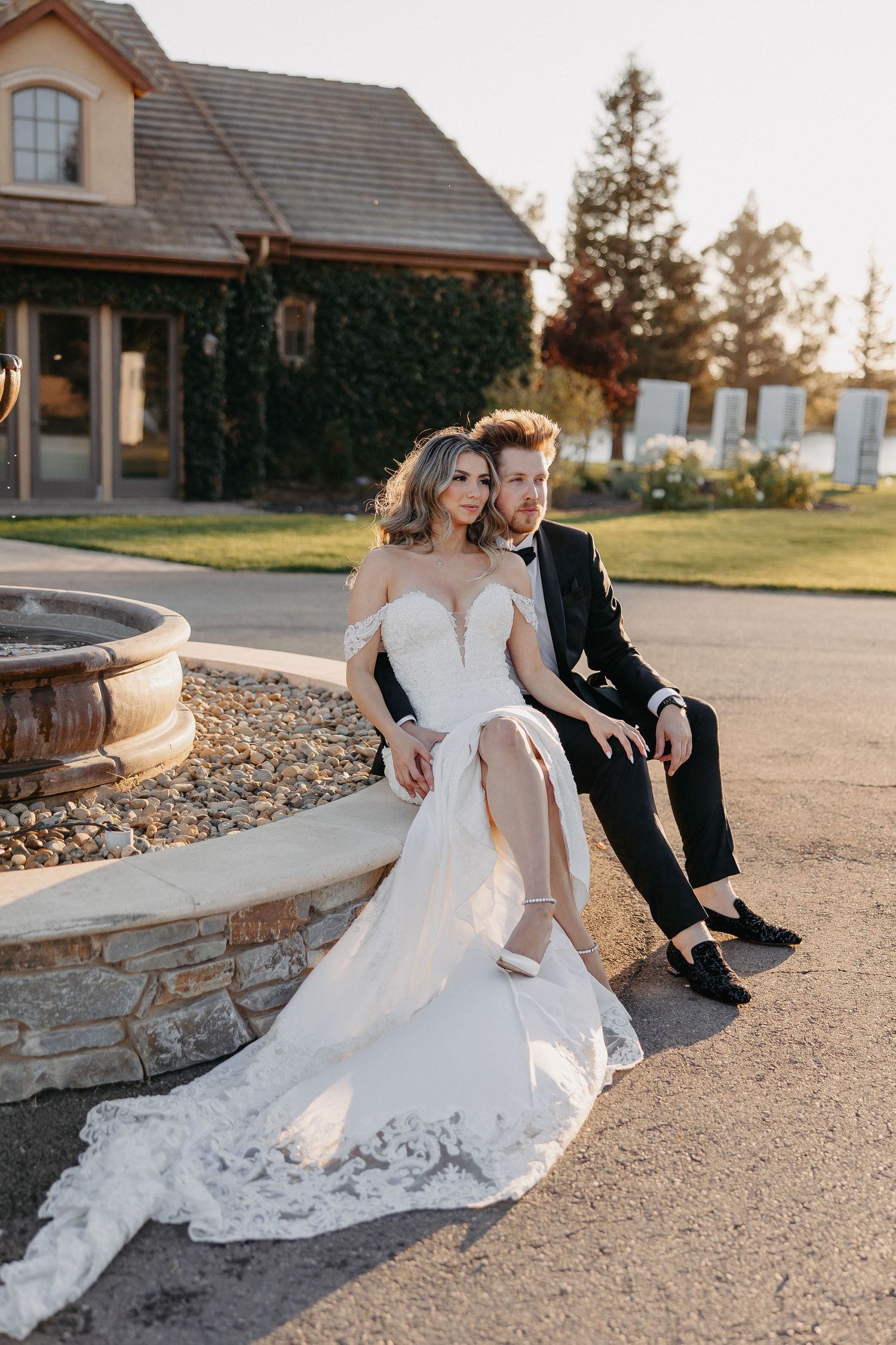 Golden hour portrait of couple sitting by stone fountain outside elegant venue.