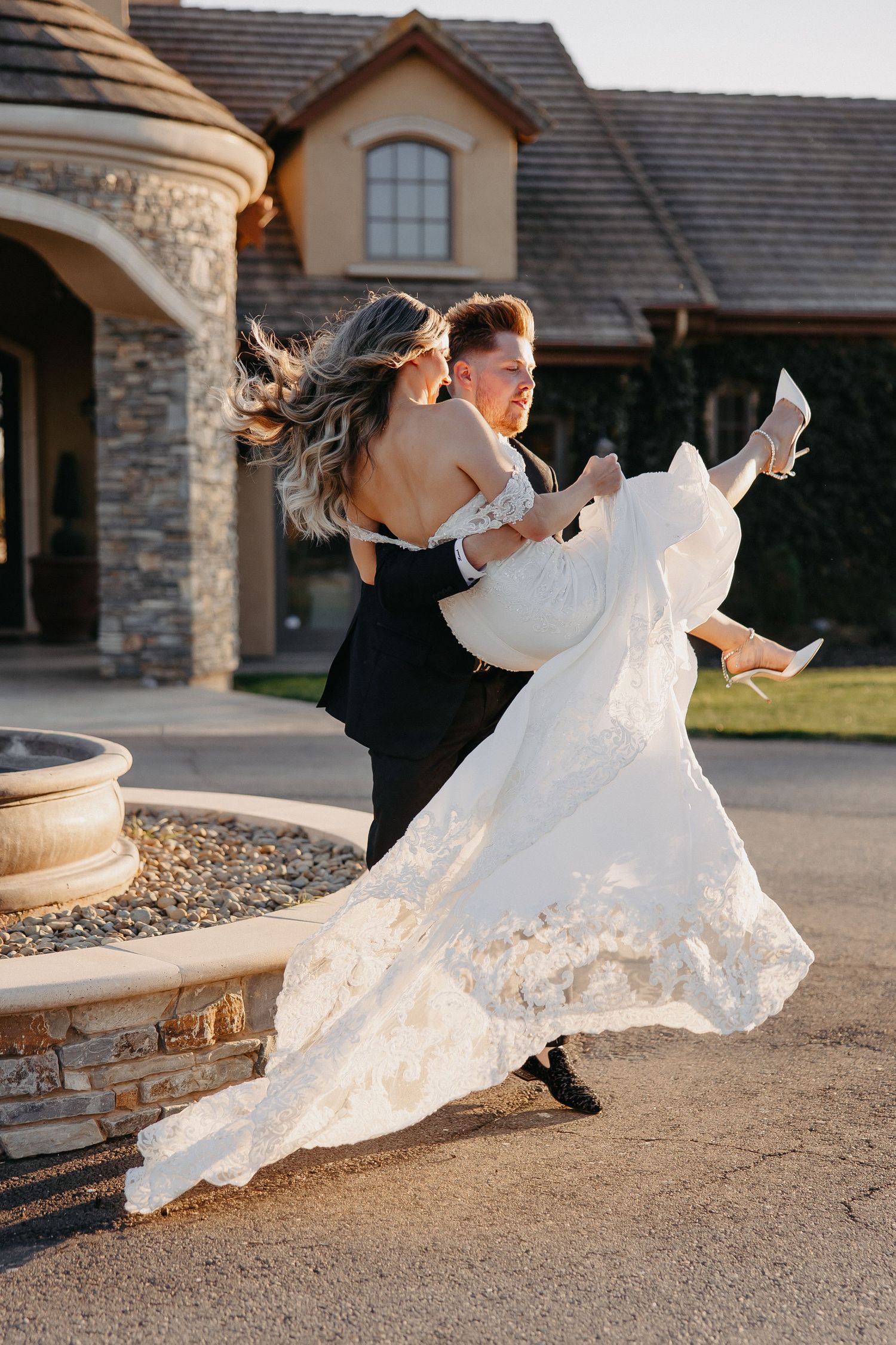 The groom lifts and spins his bride in a flowing white dress outside their venue.