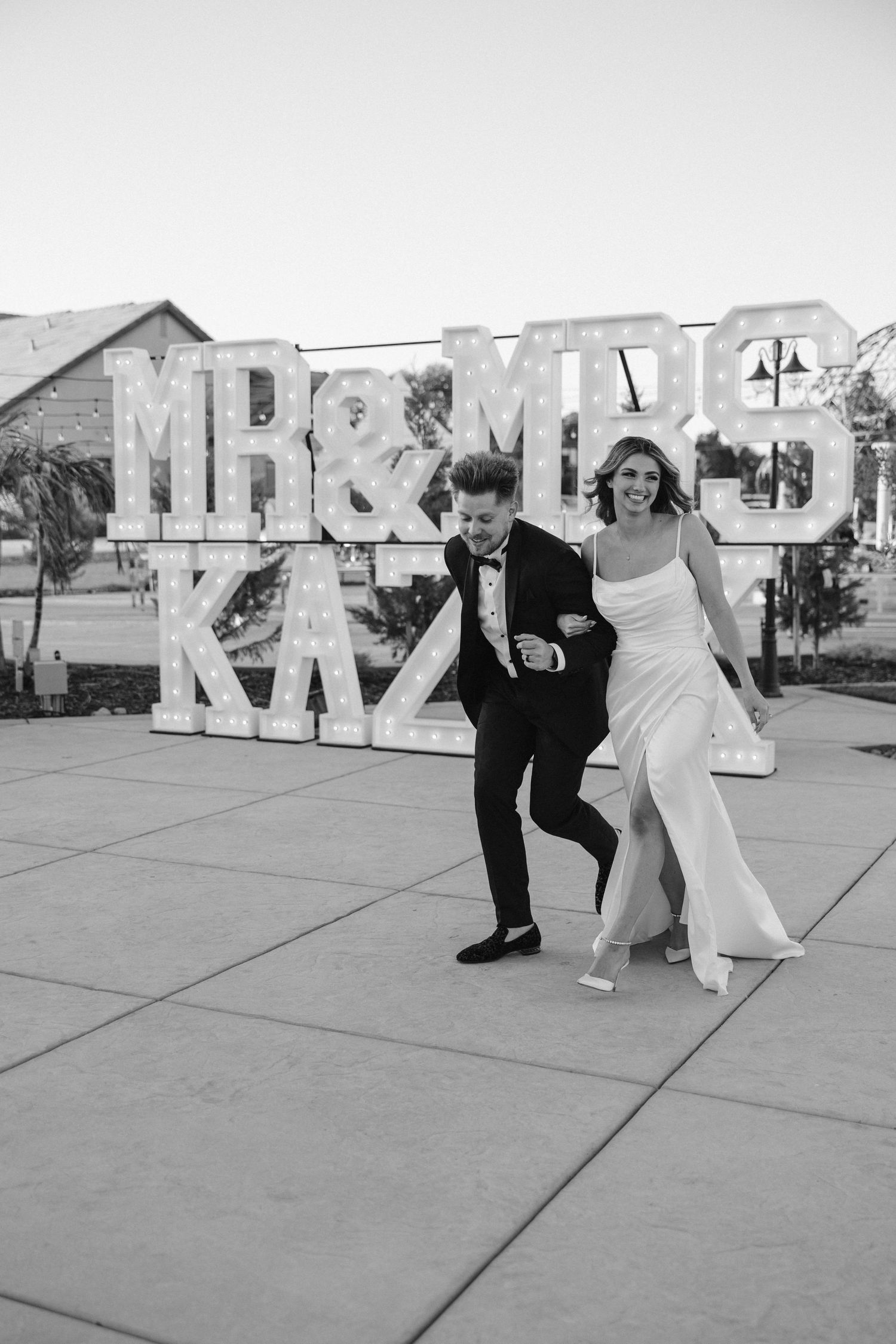 A joyful wedding couple dances and laughs in front of large marquee letters outdoors.