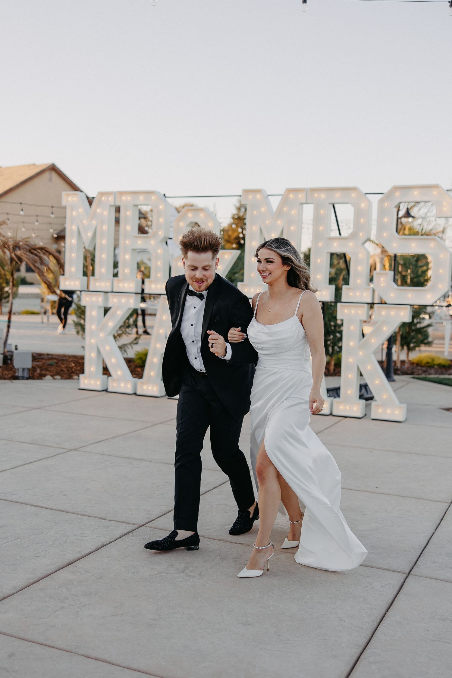 A couple in wedding attire dances playfully in front of large decorative letters.