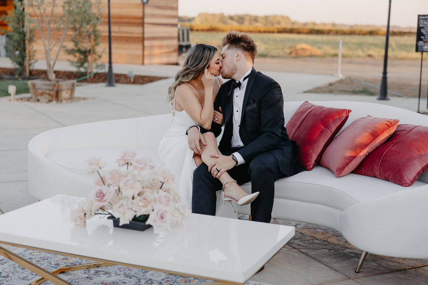 A wedding couple shares an intimate moment on a white outdoor lounge sofa with pink floral decorations.