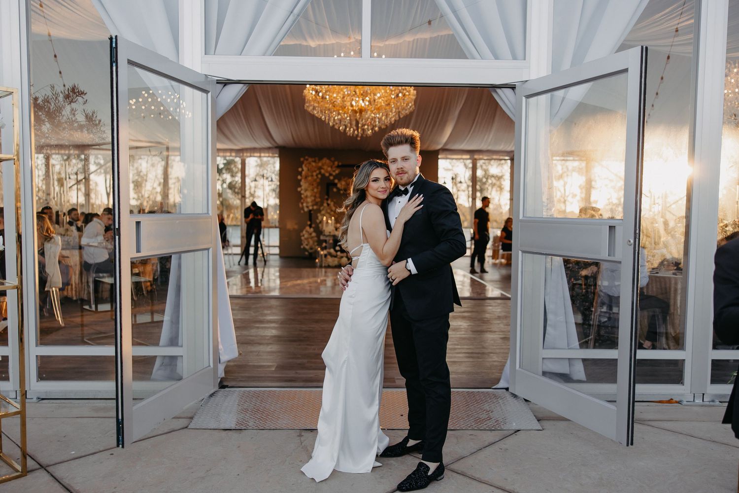 A wedding couple poses in an elegant white draped entrance with a crystal chandelier.