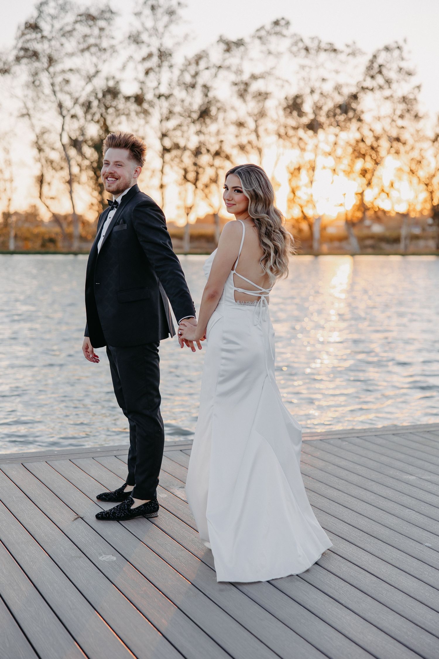 A couple holds hands on a wooden dock at sunset with trees silhouetted in the background.