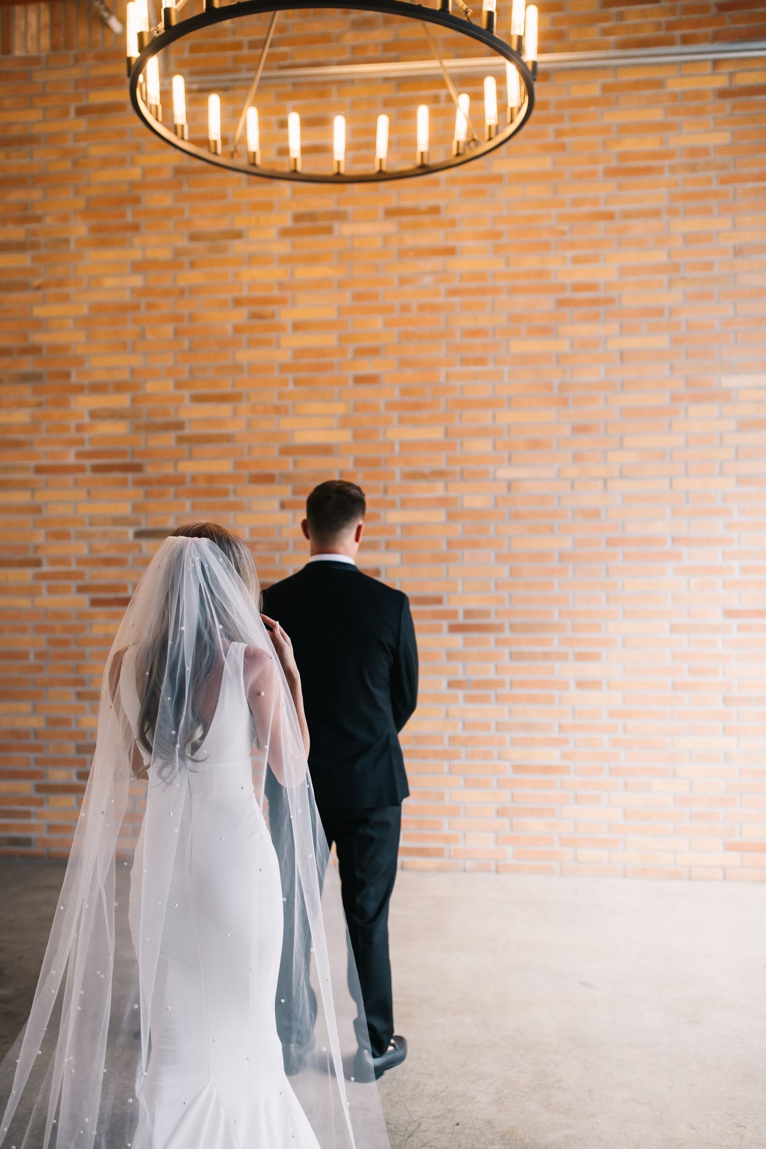 Silhouette of newlyweds under circular chandelier against brick wall in elegant venue.