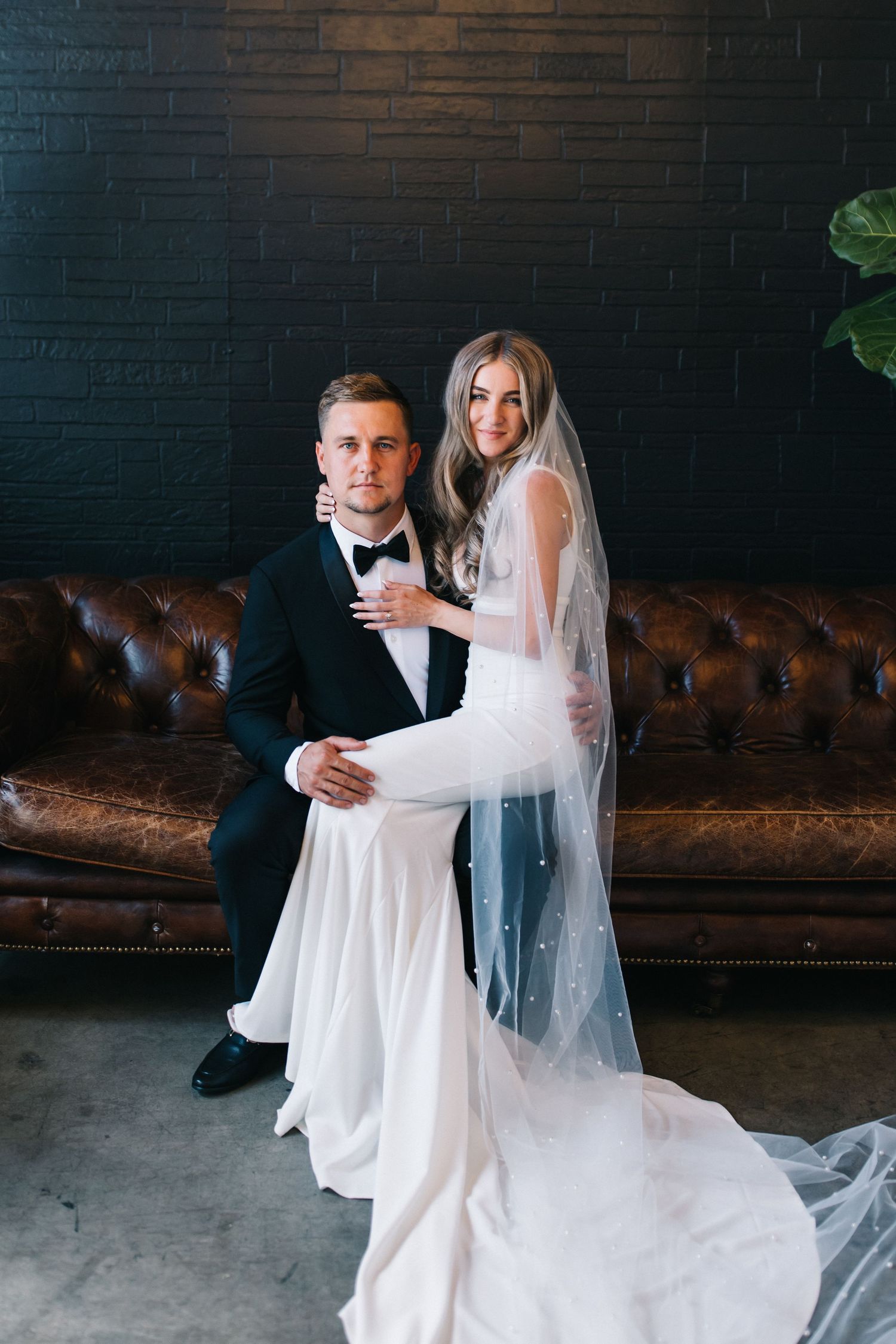 Bride and groom pose on vintage leather couch against dark brick wall.