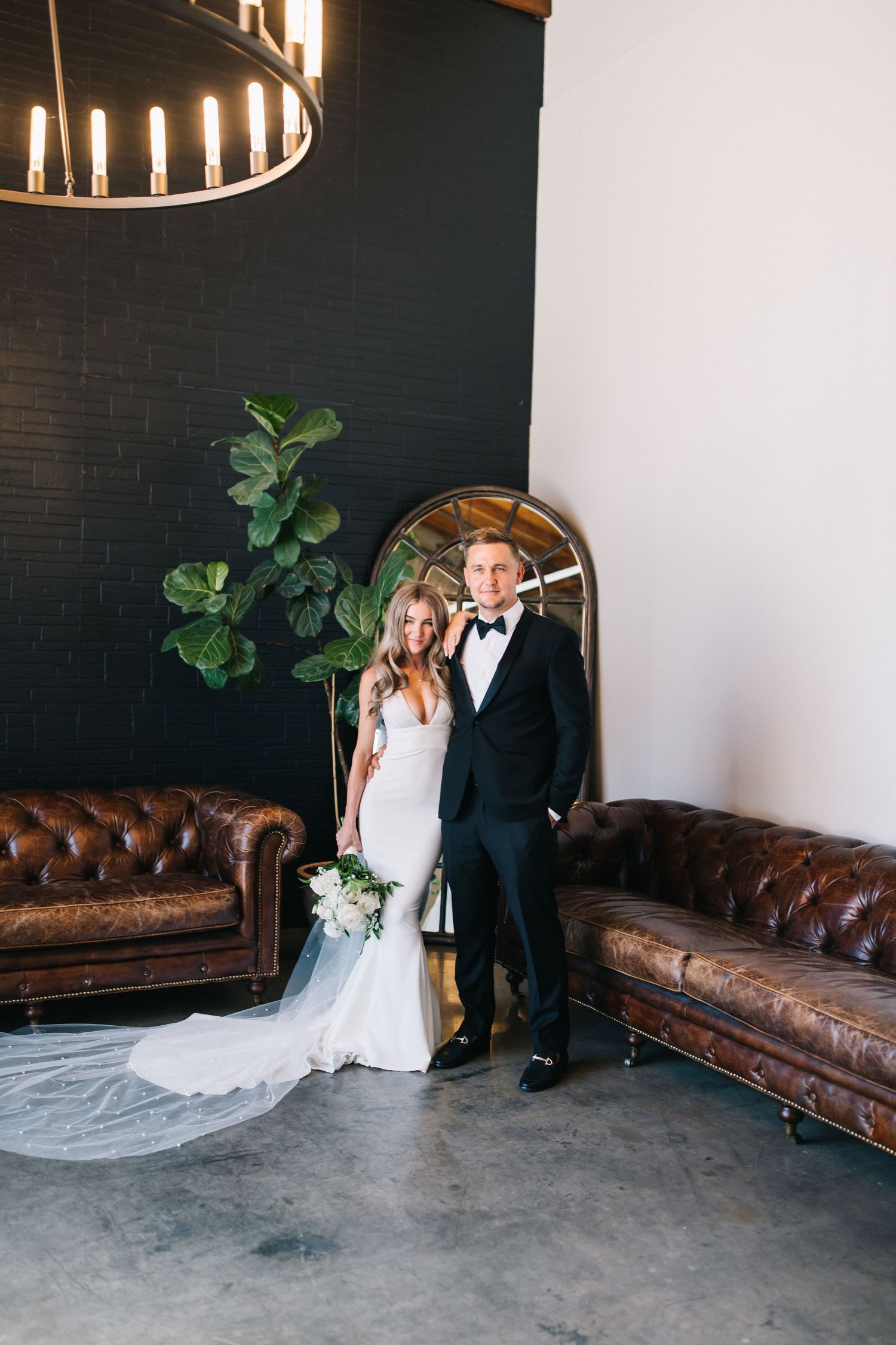 Wedding couple poses by arched mirror and leather couches in industrial chic setting.
