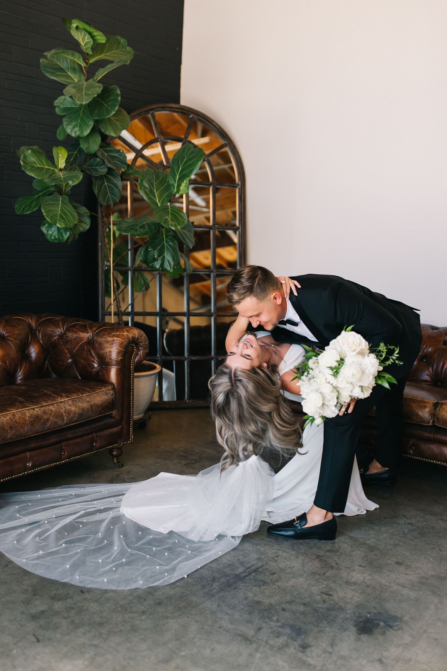 Elegant wedding dip pose with white bouquet and vintage arched mirror backdrop.