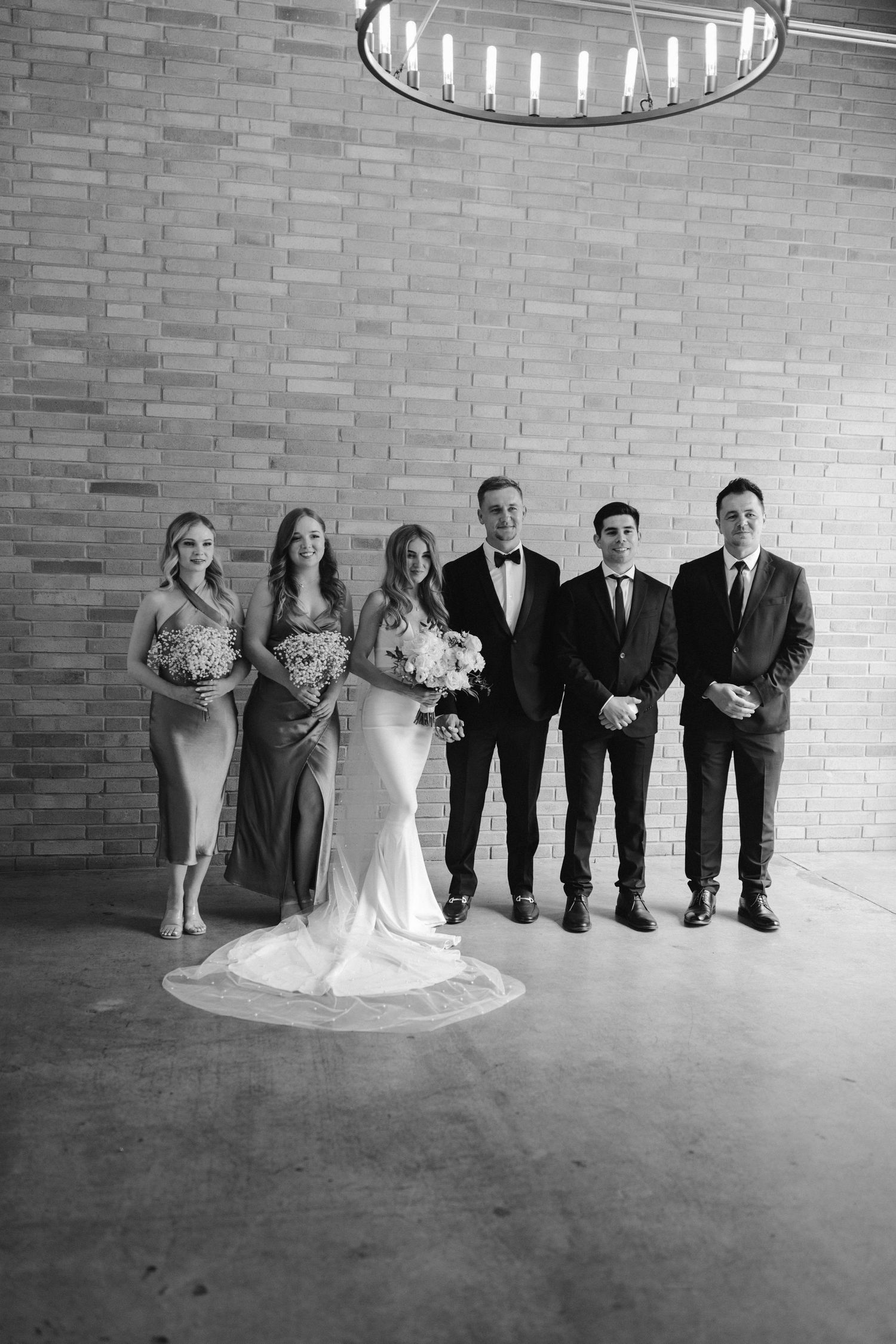 Wedding party stands posed against a textured brick wall beneath a modern circular chandelier in black and white.