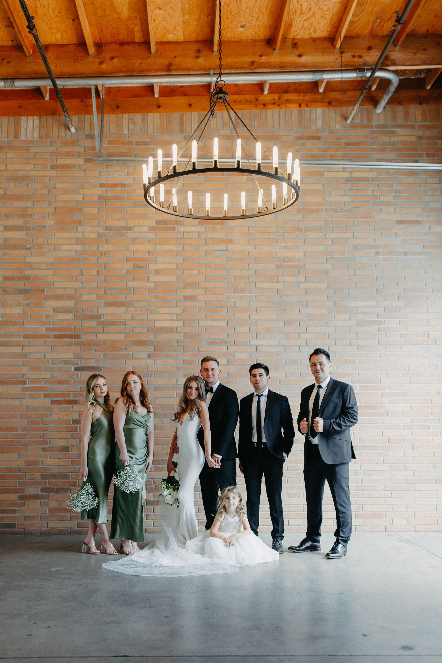 Wedding party gathers under a large ring chandelier against brick wall wearing formal attire.