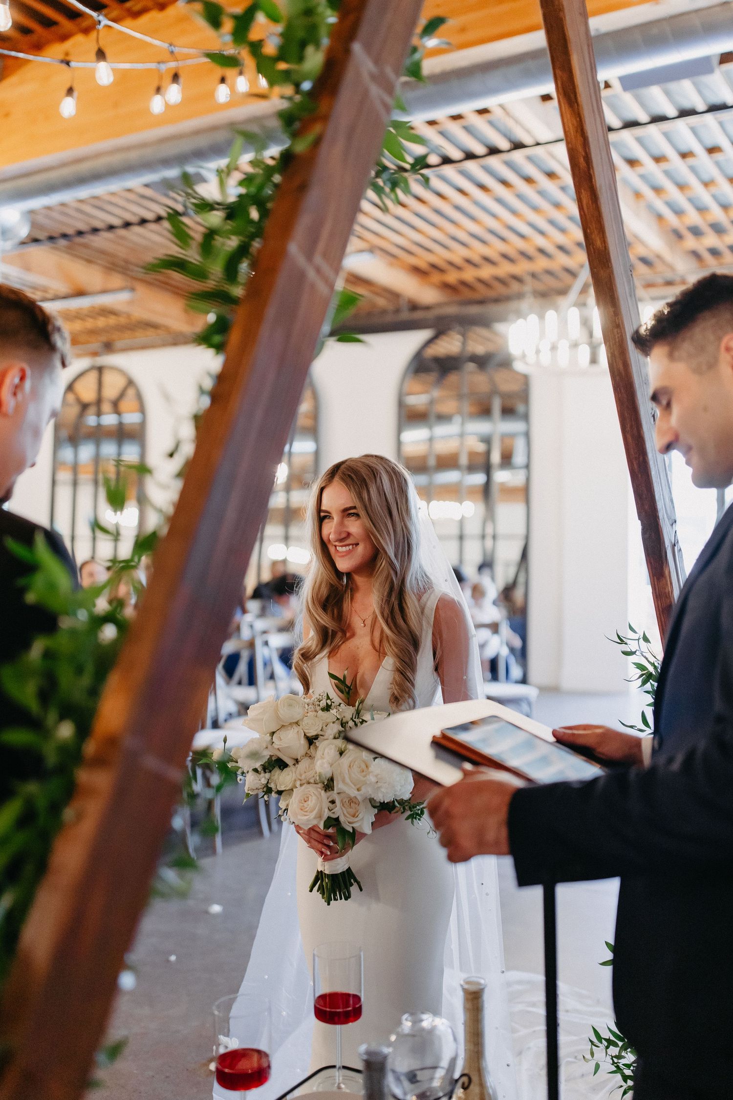Wedding ceremony takes place under wooden beams with string lights and greenery.