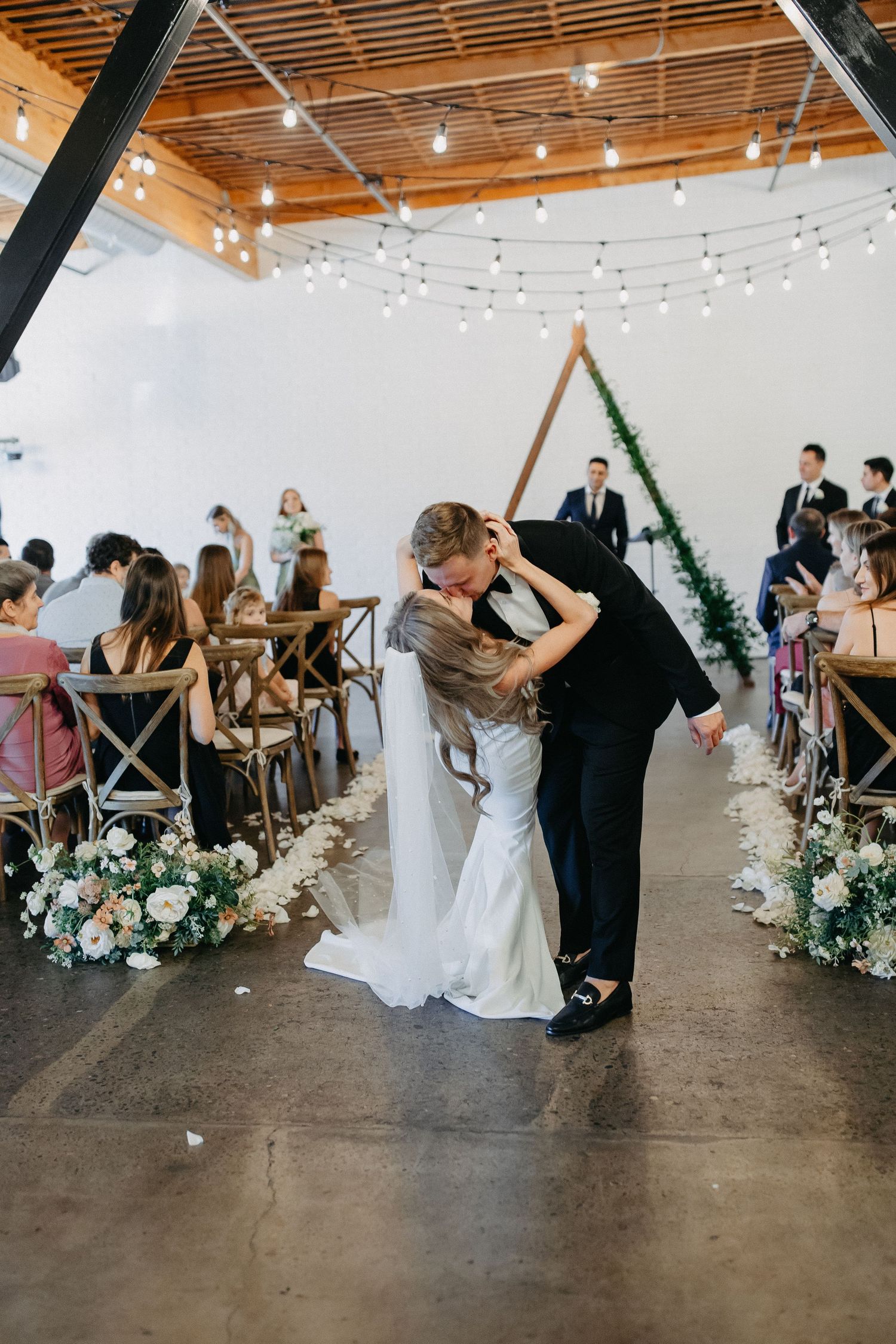 Wedding ceremony in modern white venue with string lights and floral aisle decorations.