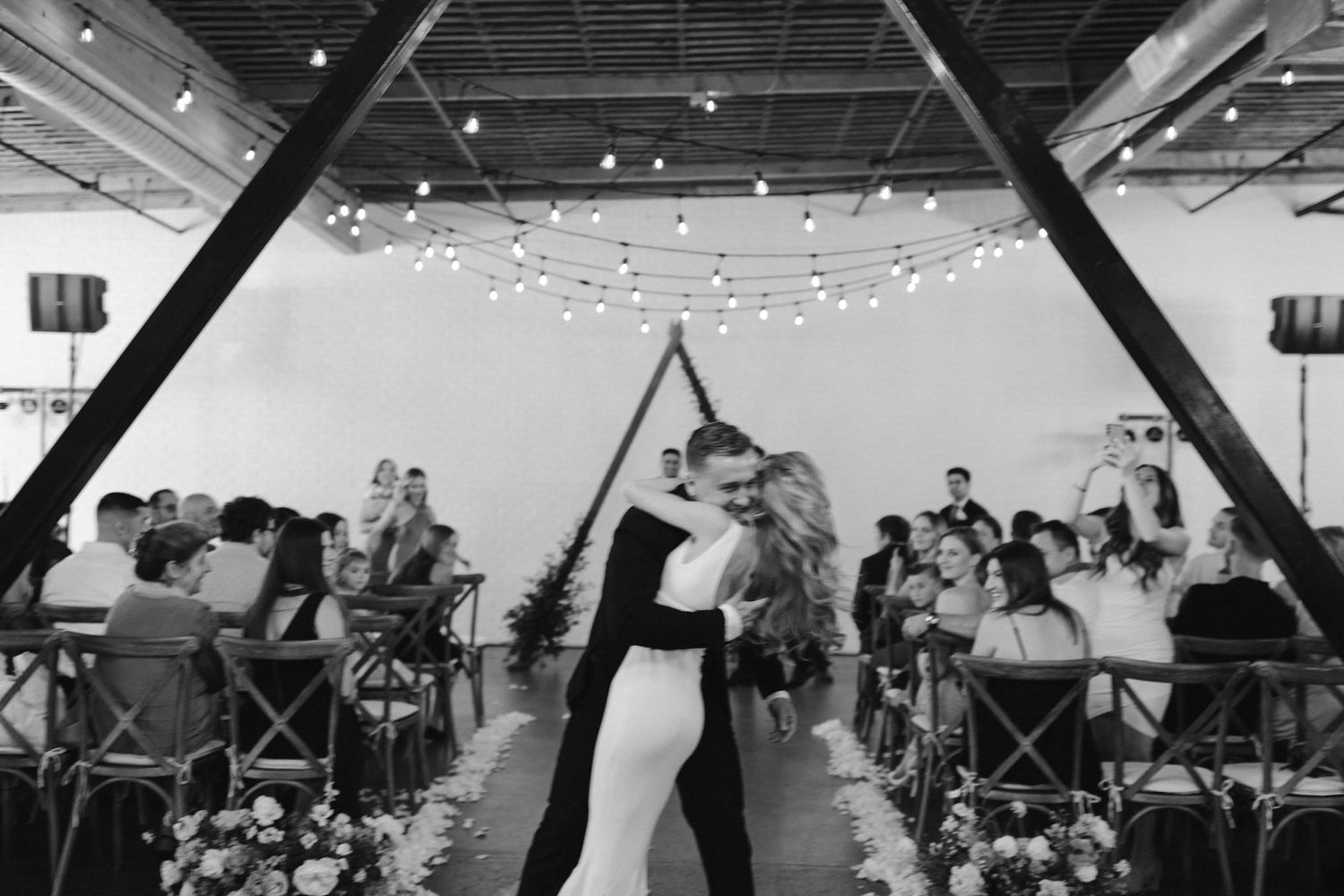 Black and white wedding ceremony shot through geometric frame with string lights overhead.