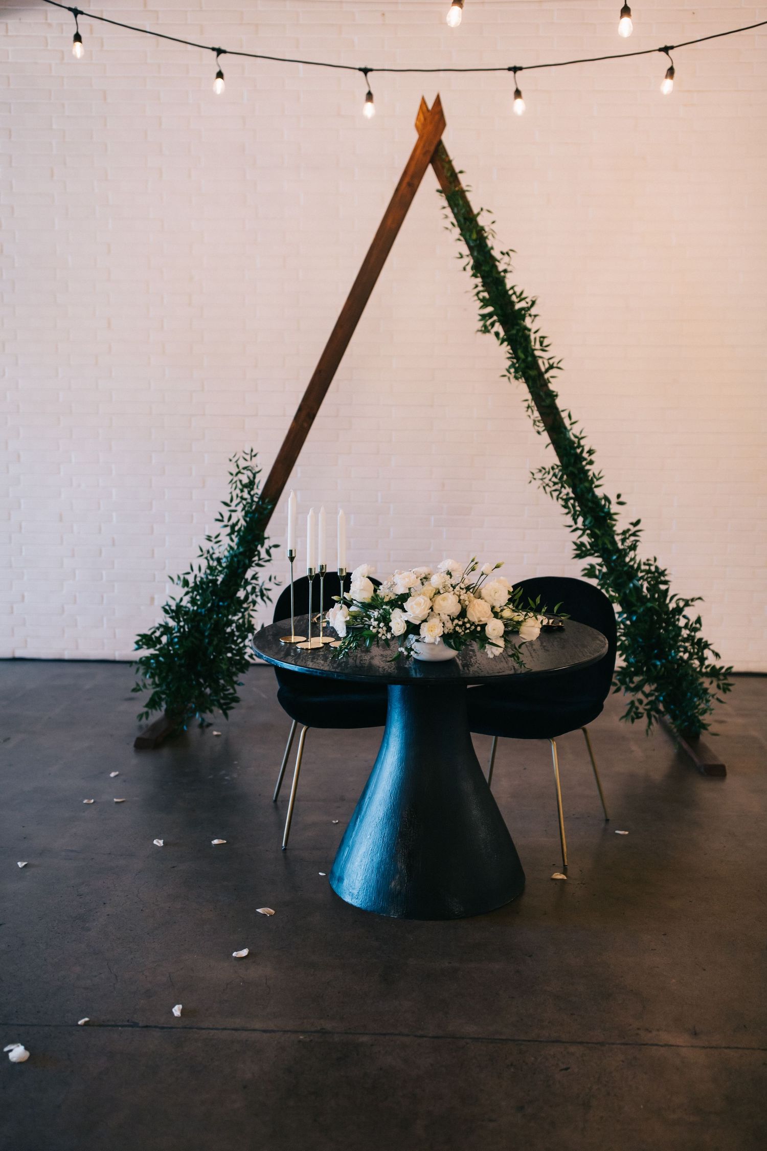 Modern wedding table setup with triangular wooden arch adorned with greenery and floral centerpiece on black pedestal table.
