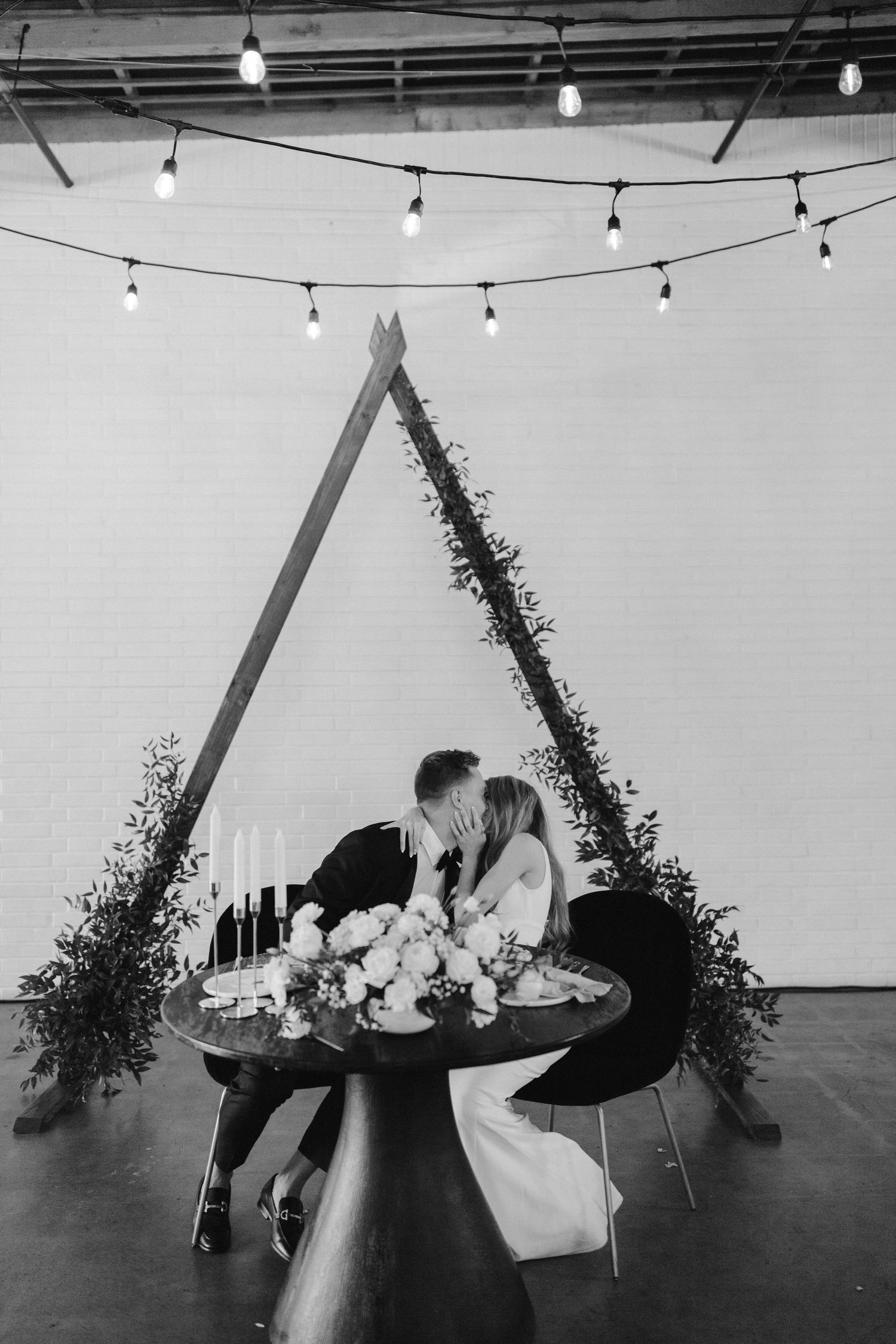 Romantic black and white photo of couple sharing intimate moment under triangle arch with string lights.