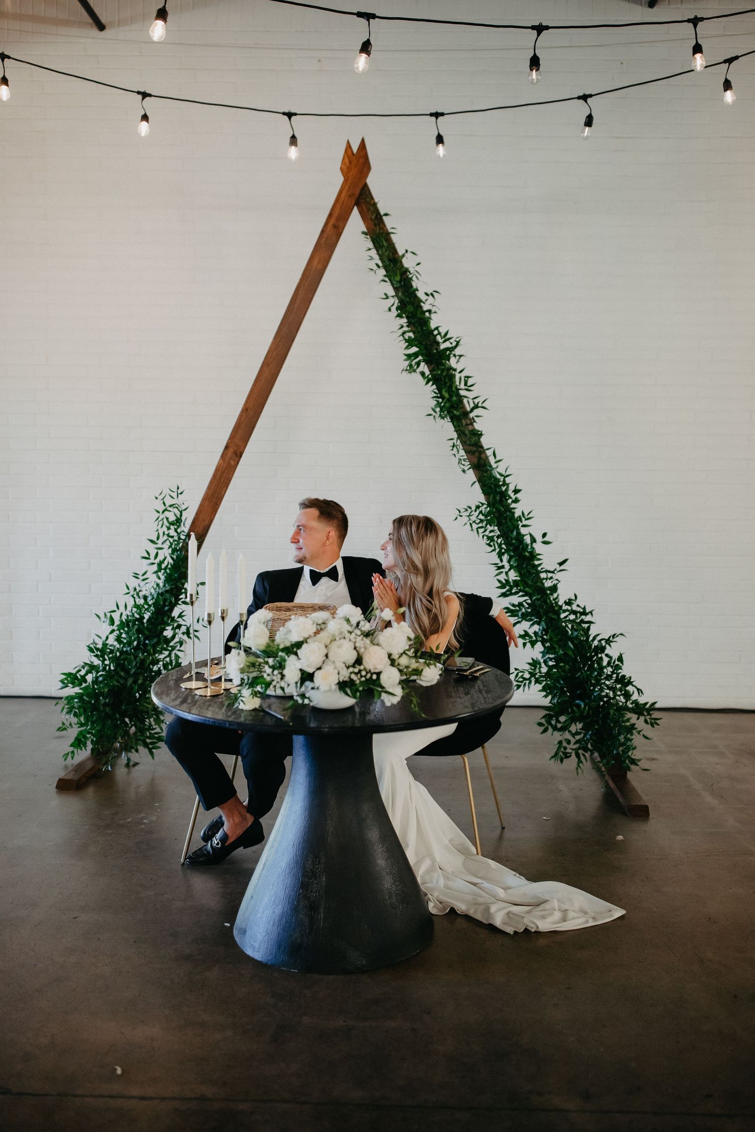 Newlyweds seated at modern pedestal table under wooden triangle arch with greenery.