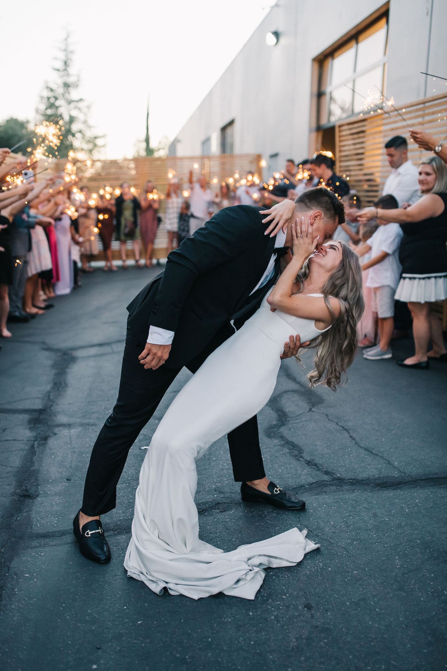 Dramatic wedding dip pose captured during sunset with guests holding sparklers in background.