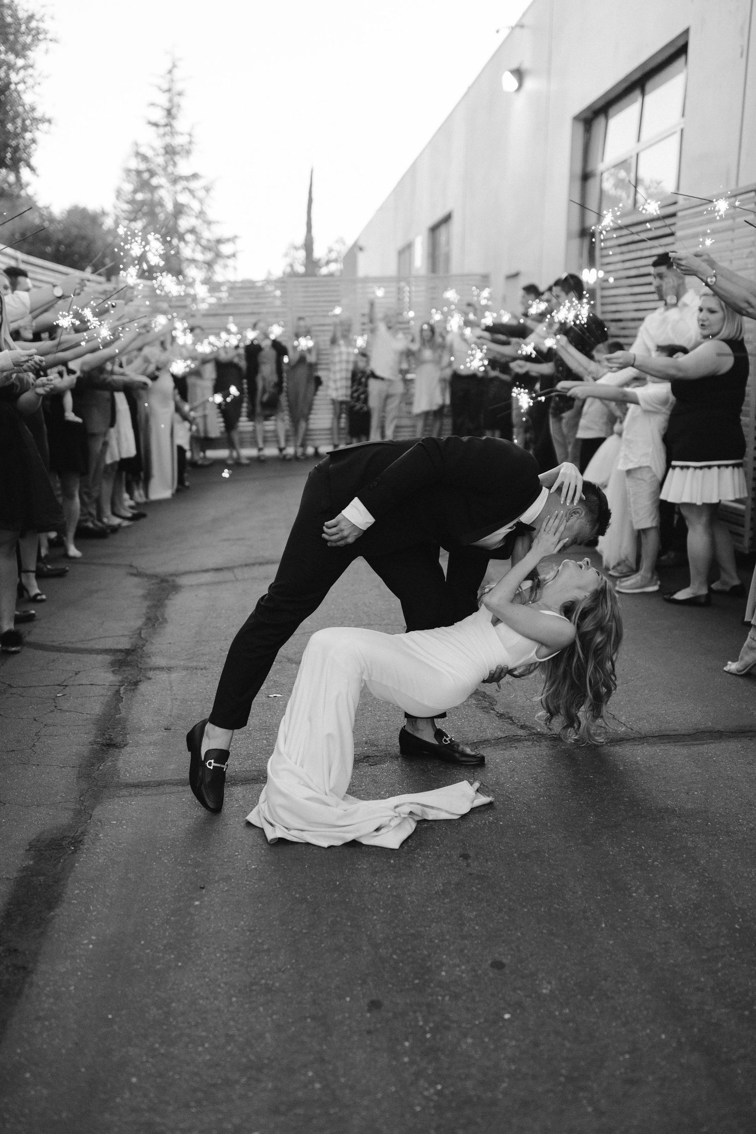 Black and white capture of dramatic wedding dip kiss with sparkler-holding guests in background.