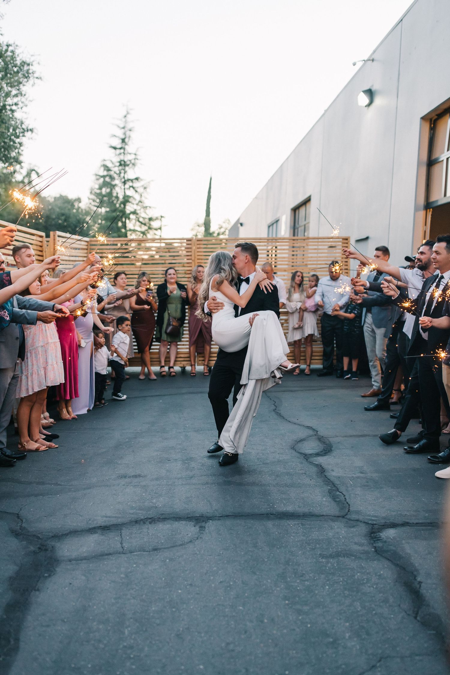 Newlyweds dancing outdoors surrounded by guests holding sparklers at sunset reception.