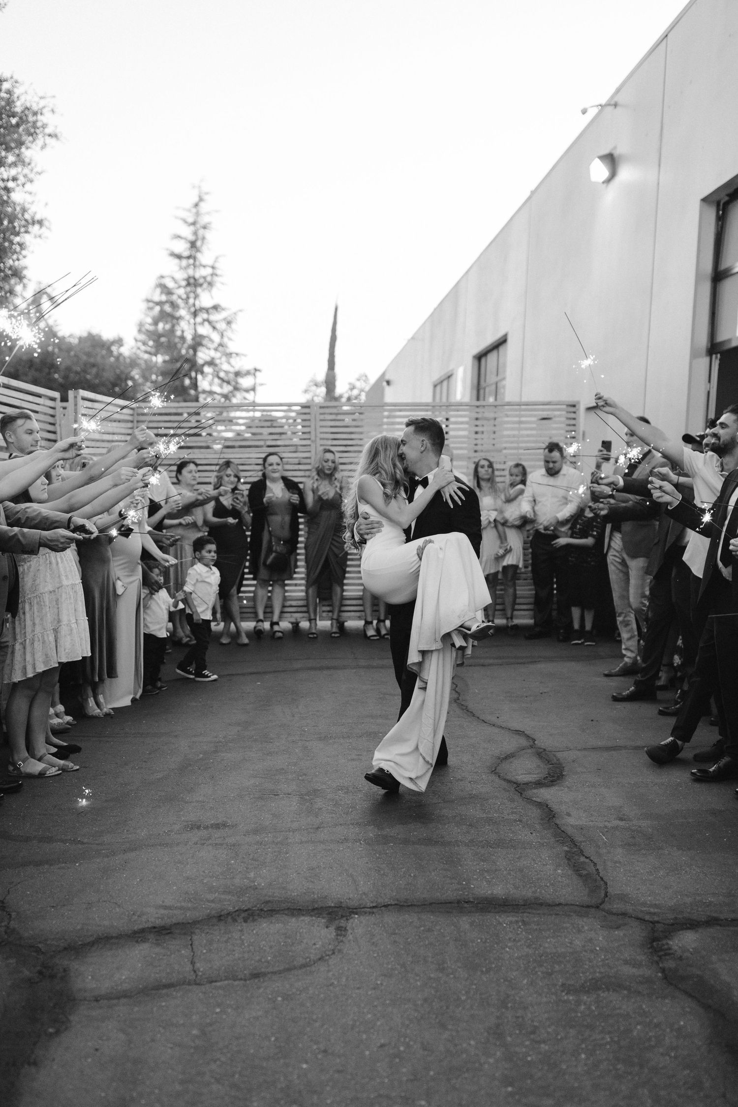 Wedding guests cheer and wave sparklers as couple shares romantic dance during outdoor evening celebration.