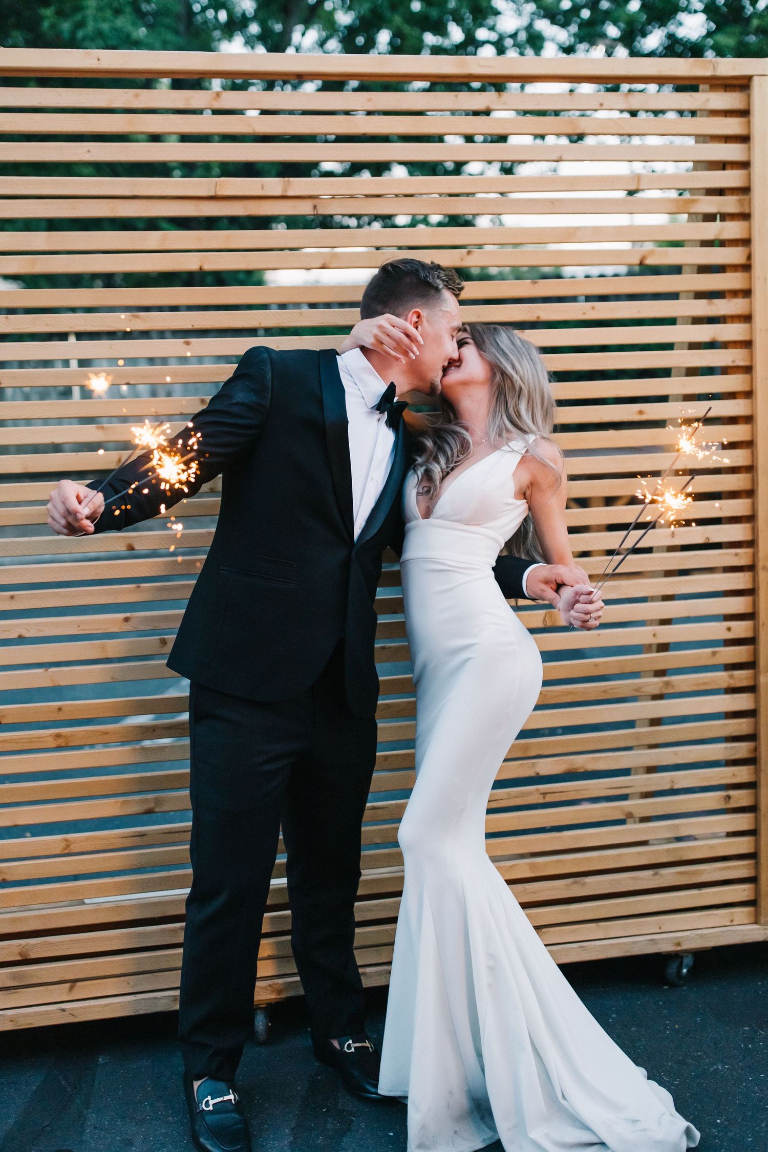 Couple in formal attire shares romantic moment with sparklers against wooden wall backdrop.