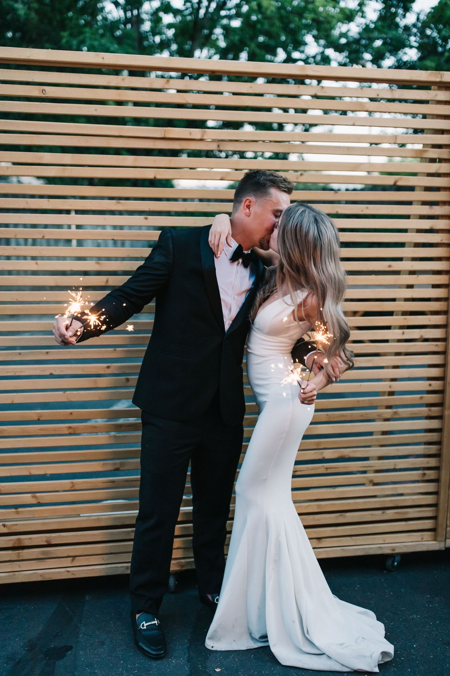 Newlyweds hold sparklers while sharing intimate moment against modern wooden wall.