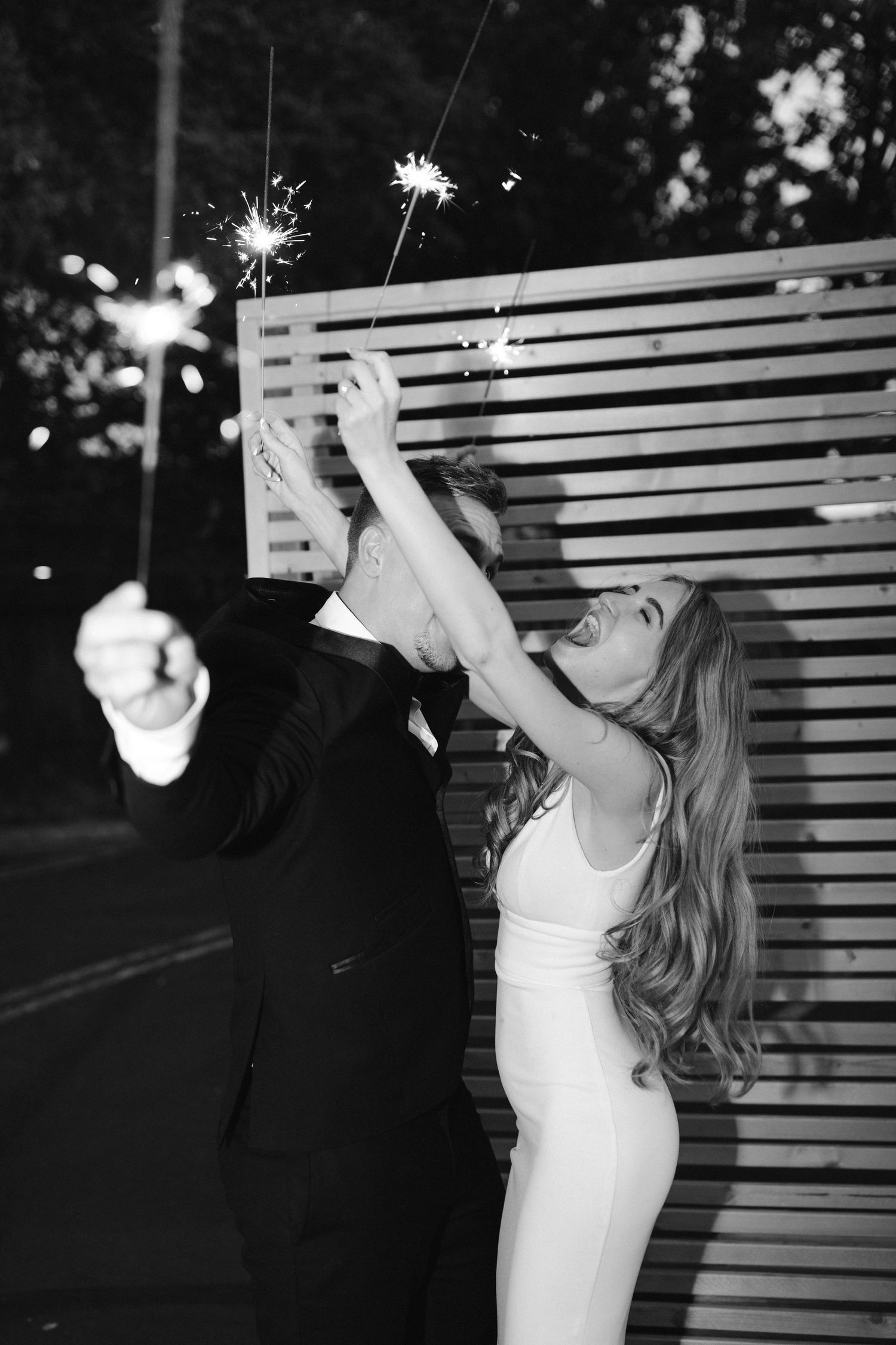 Couple celebrates with sparklers against wooden slat wall in black and white photo.