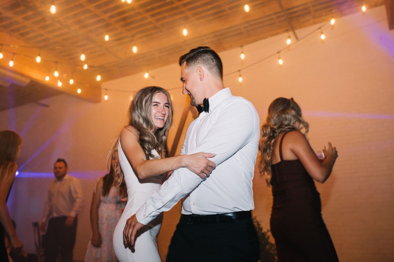 Couple sharing a lively dance moment under warm string lights at wedding reception.
