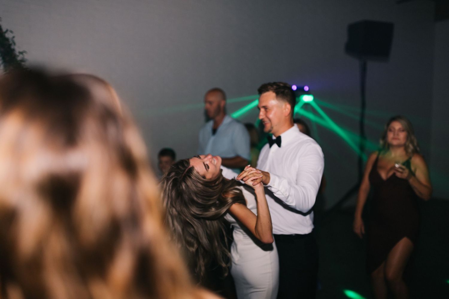 A couple sharing a romantic dance moment on a dimly lit dance floor surrounded by guests at a celebration.