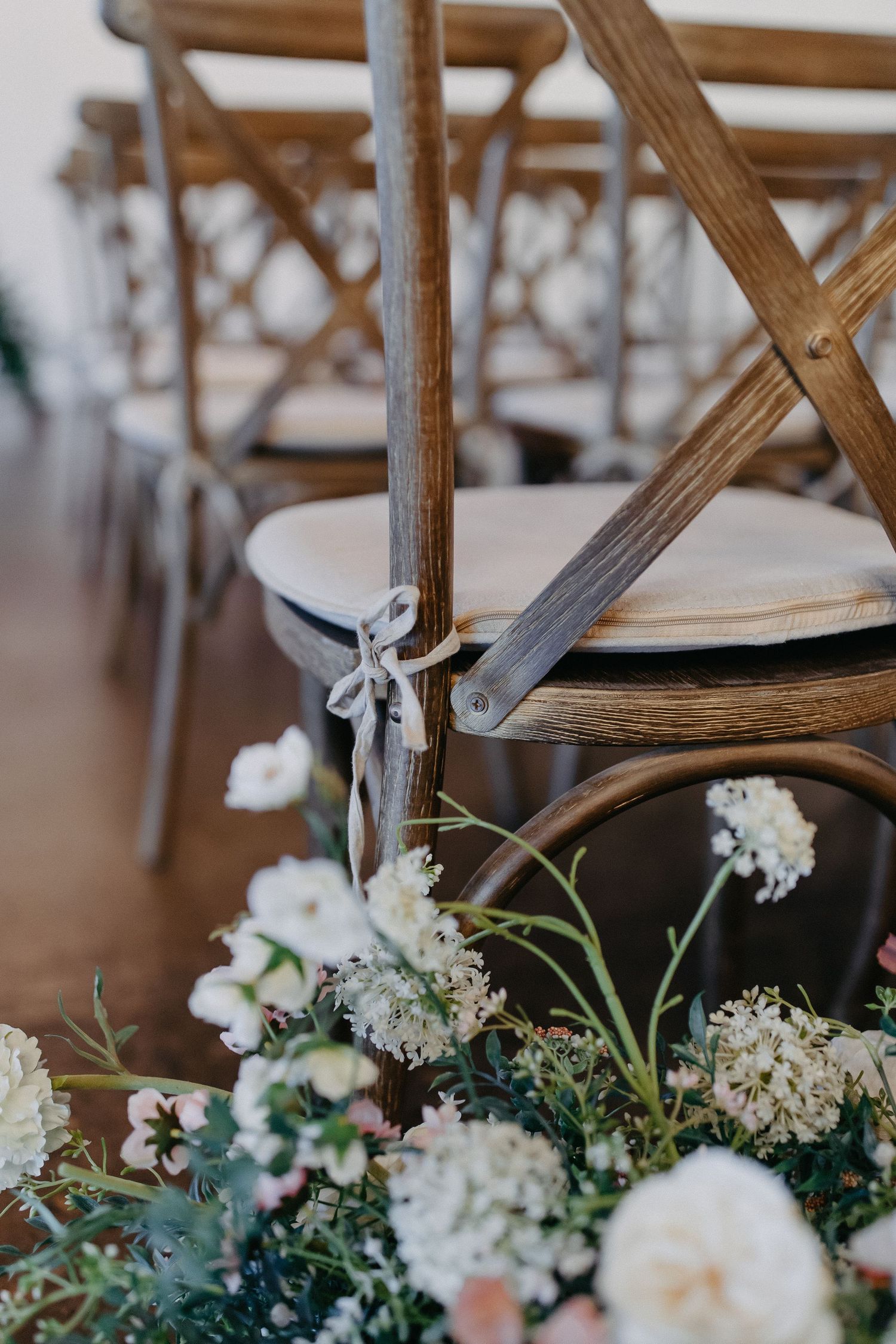 White floral arrangements decorating wooden cross-back chairs at an event.