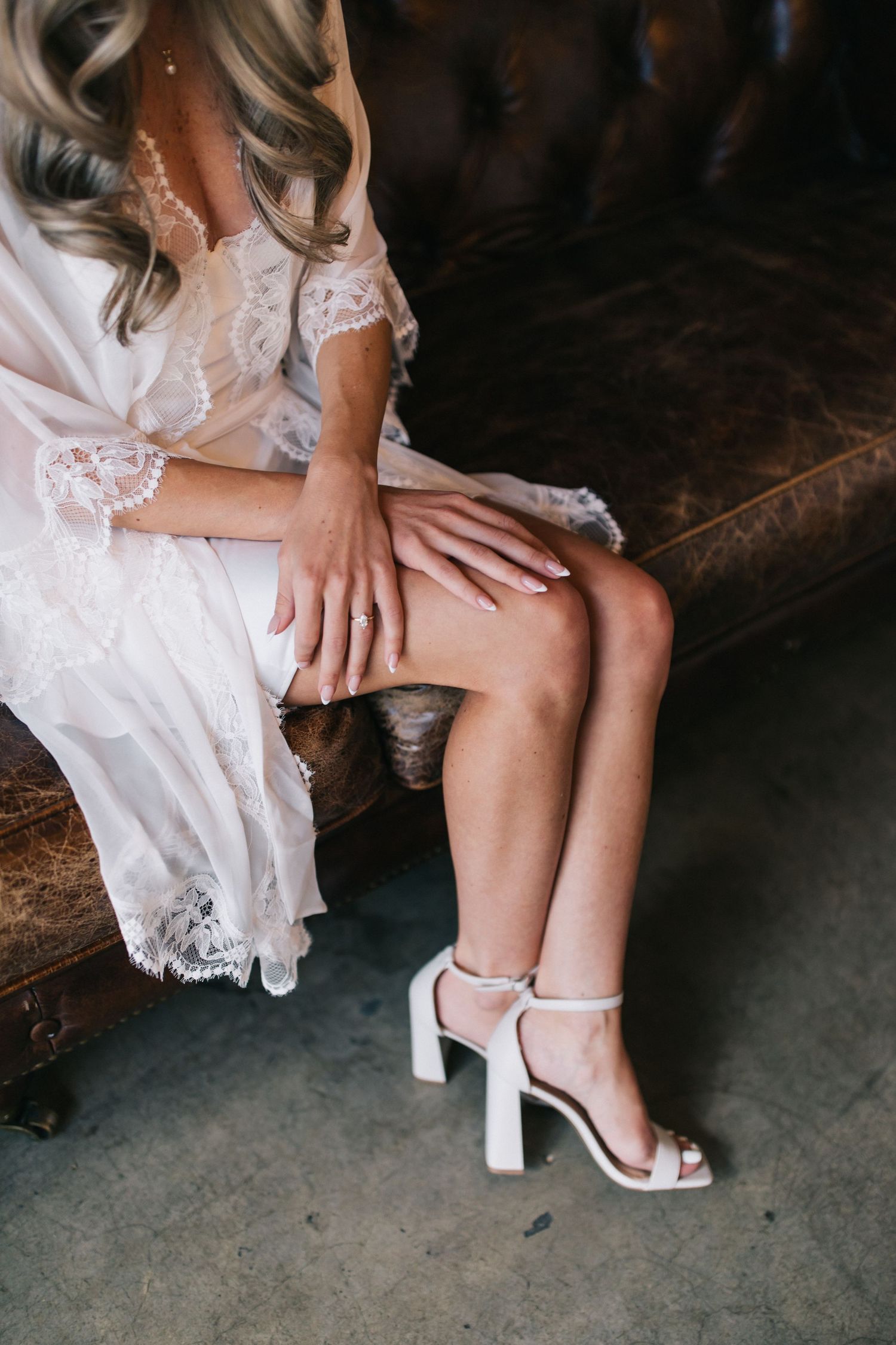 Close-up of white heels and lace dress hem on leather sofa.